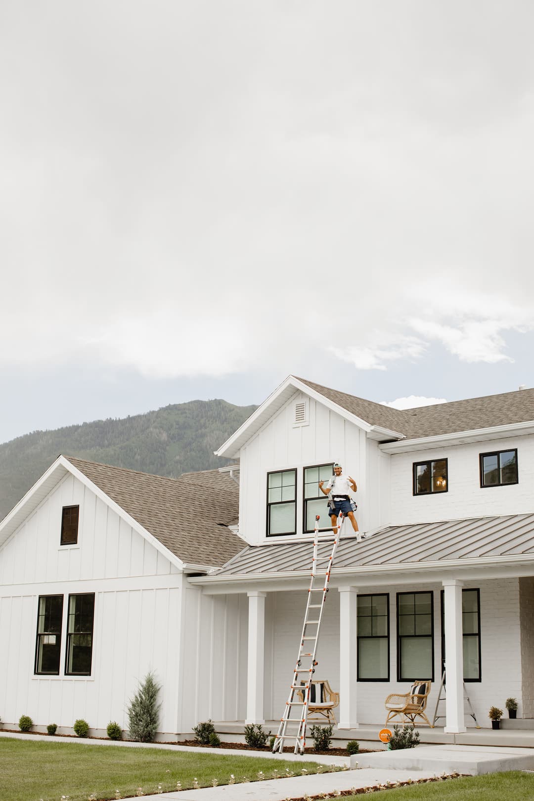 Worker on a ladder performing maintenance on a modern white house with mountain backdrop.