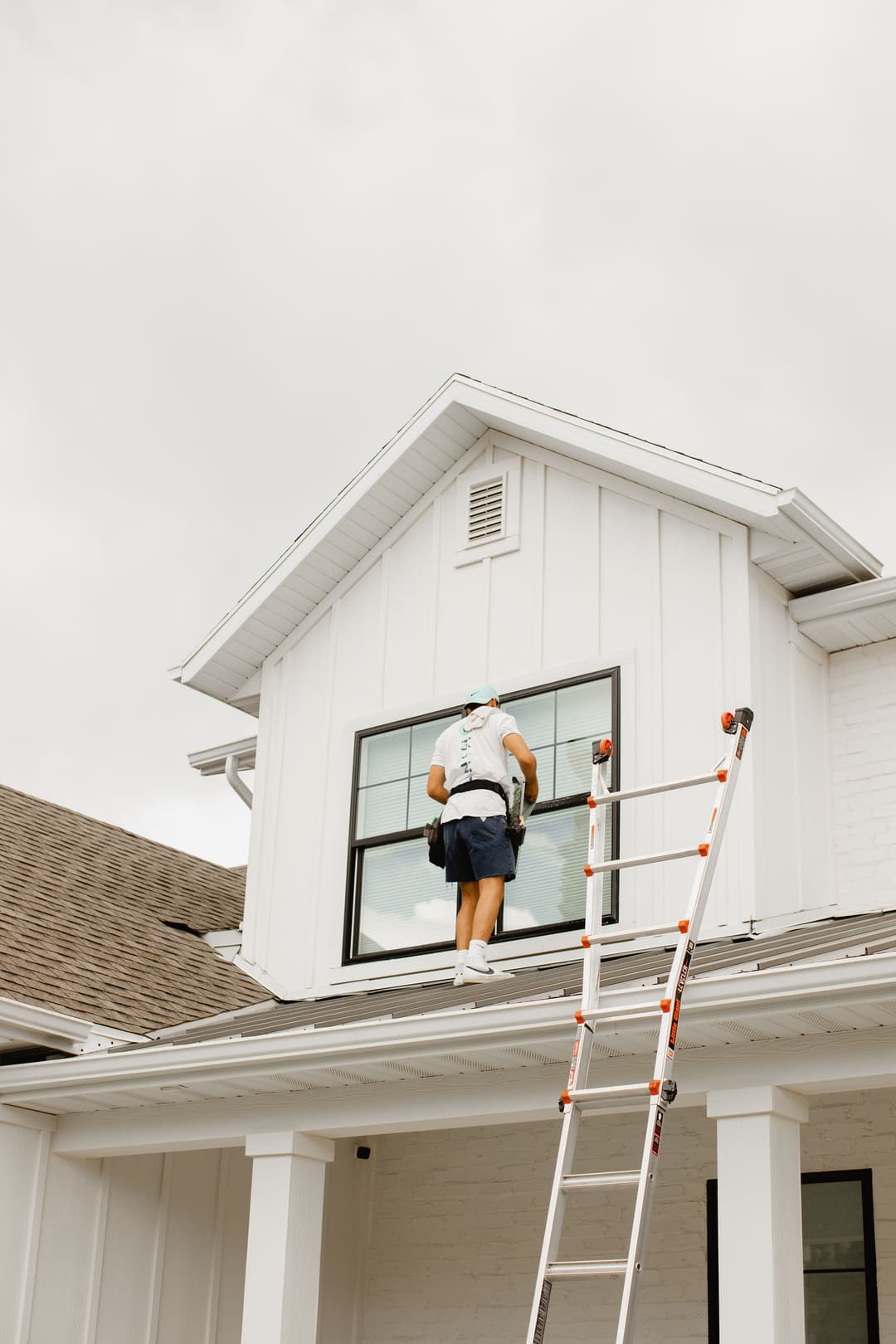 Man installing or cleaning a window on a modern house using a ladder. Overcast sky.