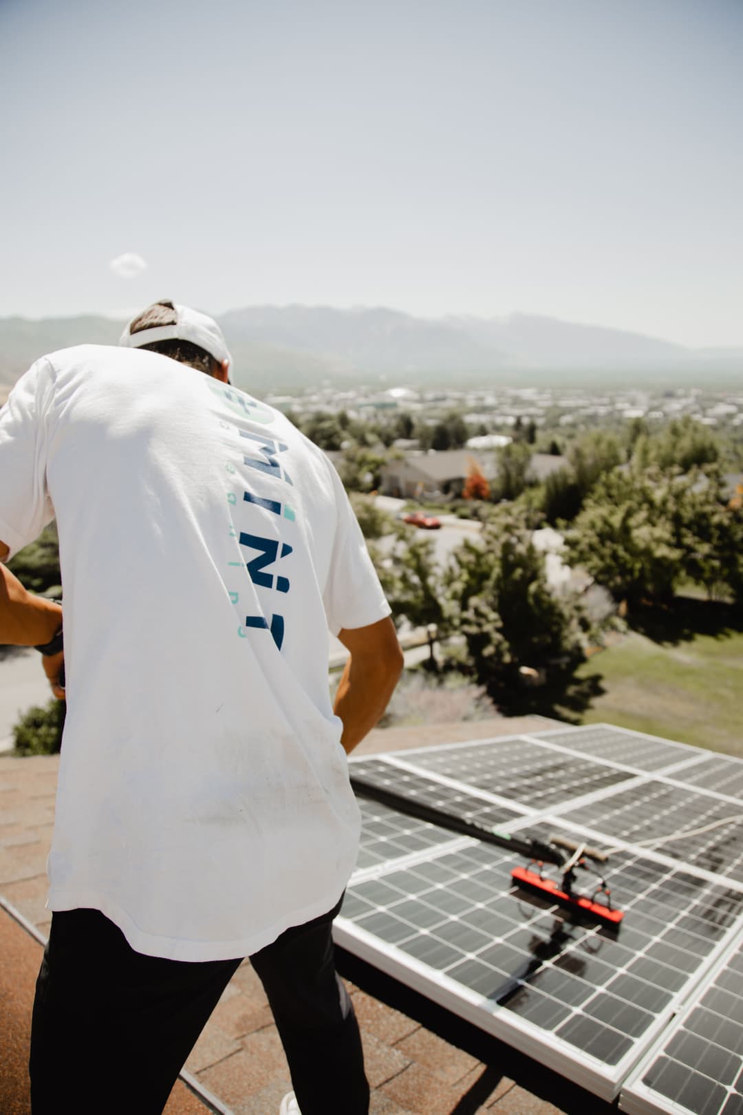Worker installing solar panels on a rooftop with mountains and cityscape in the background.