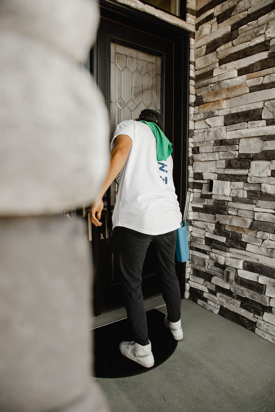 Person in a white shirt knocking on a stone house door with a green scarf.
