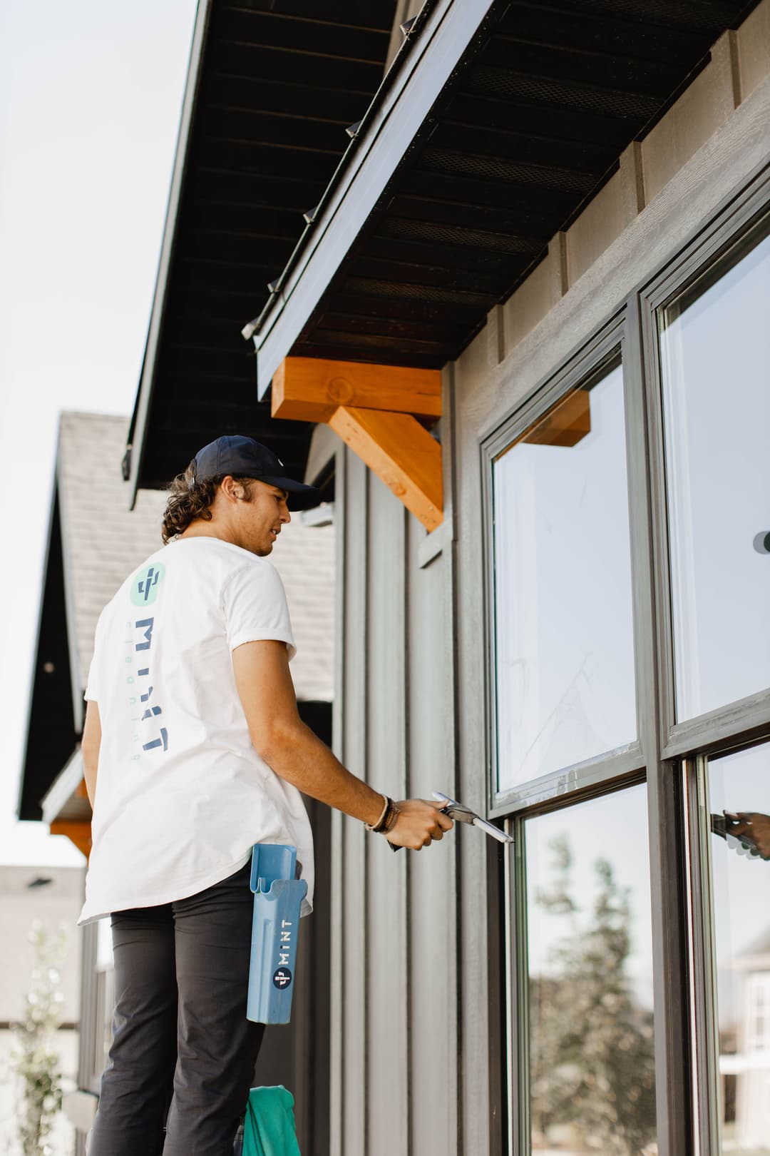 Man cleaning windows of a modern home with a squeegee, wearing a branded white t-shirt.