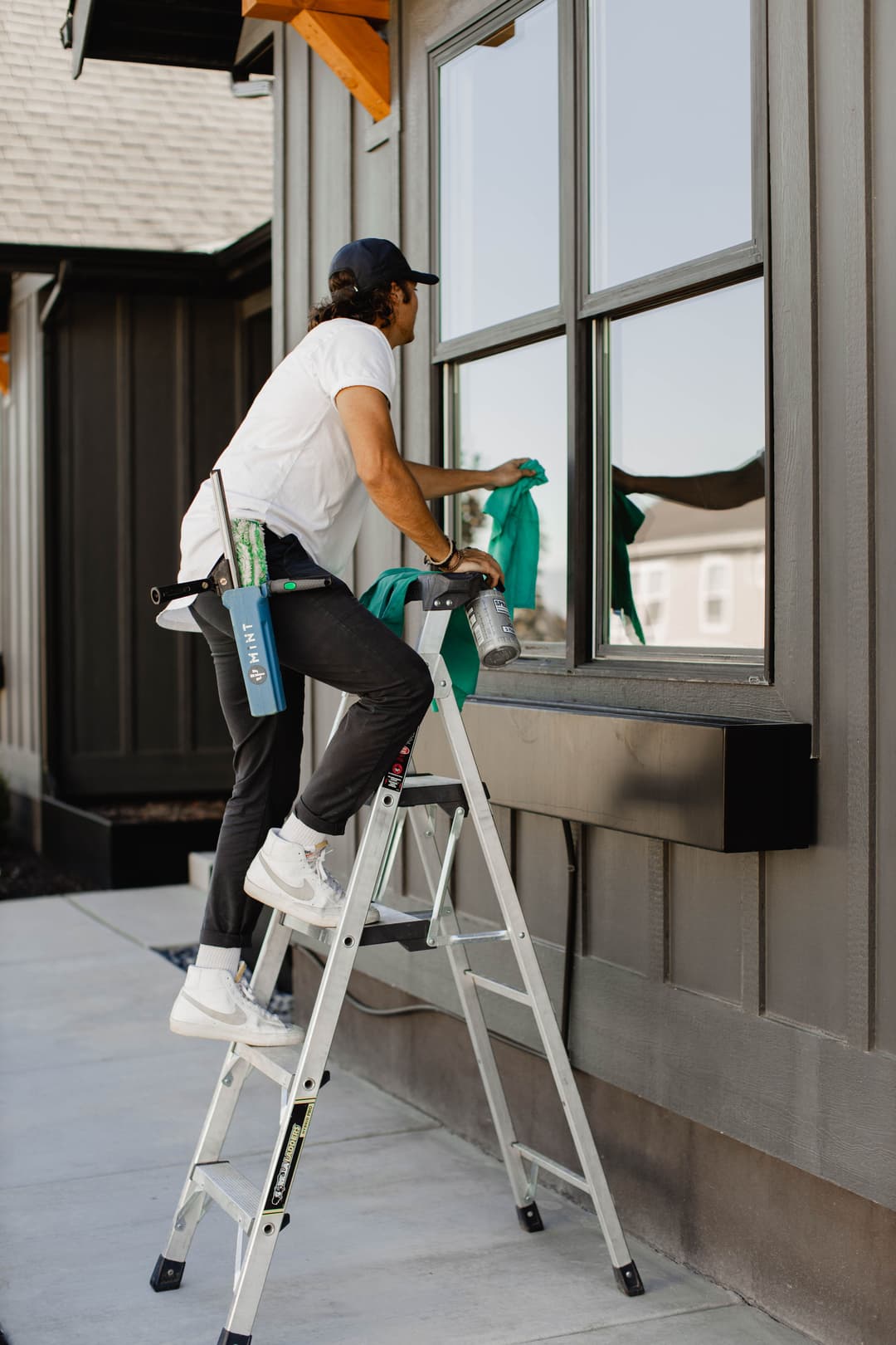 Man cleaning a window while standing on a ladder outside a modern house.