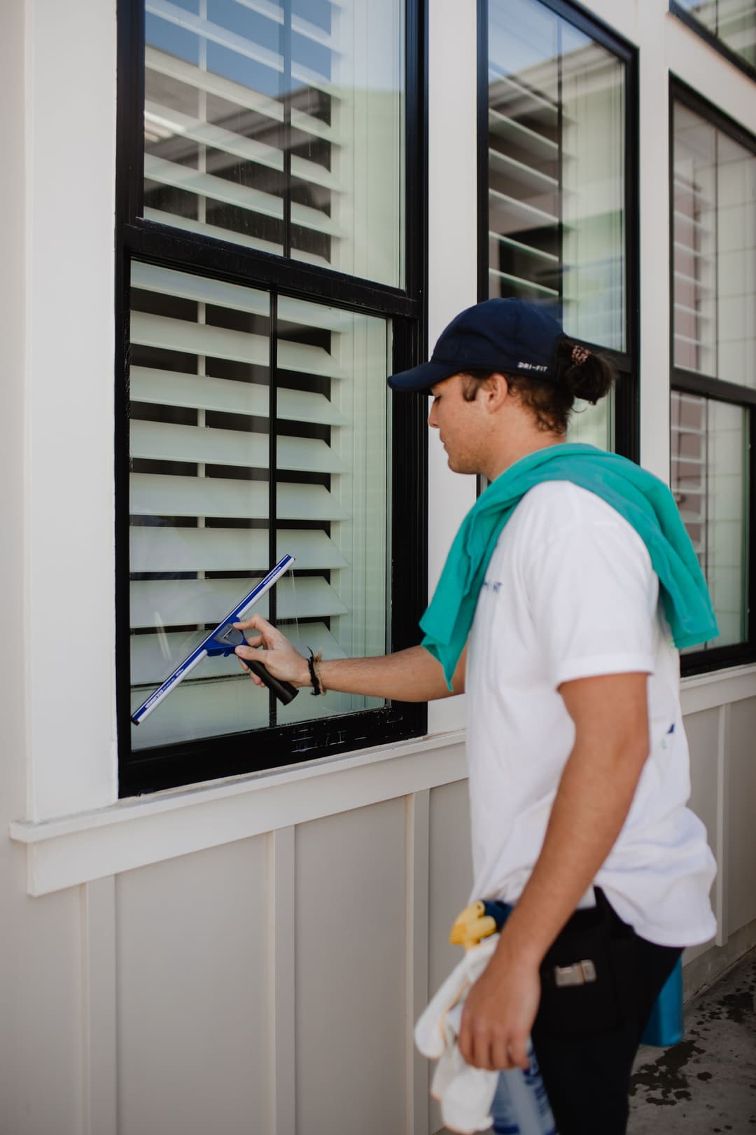 Man cleaning a window with a squeegee, wearing a cap and green towel, in a bright setting.