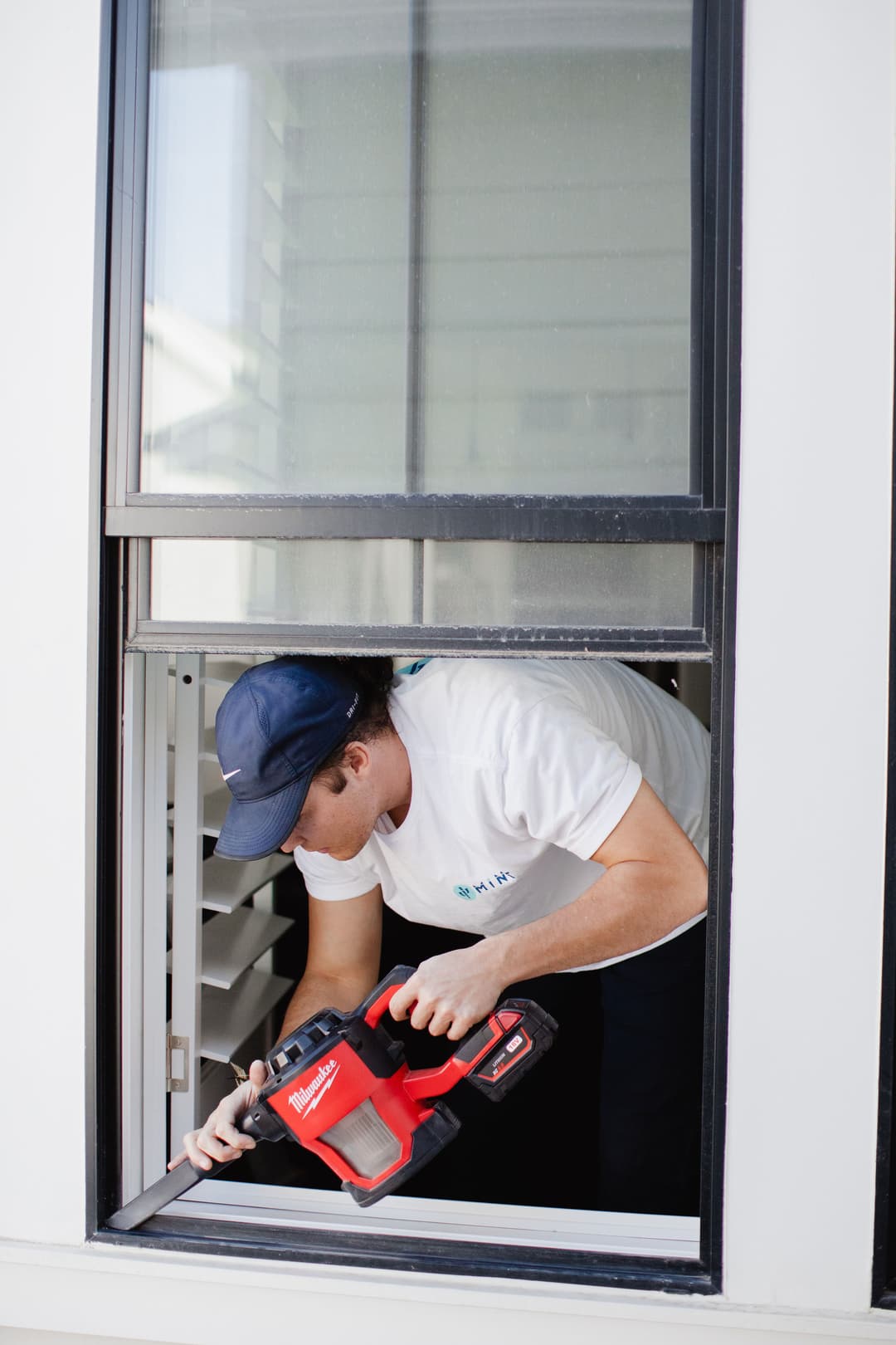 Technician using a red cordless vacuum cleaner to clean window shutters indoors.