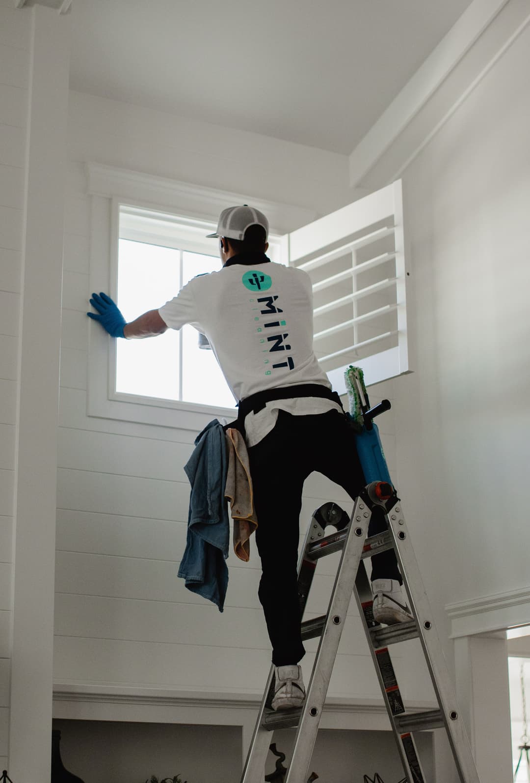 Person cleaning a window on a ladder indoors, wearing gloves and a branded shirt.