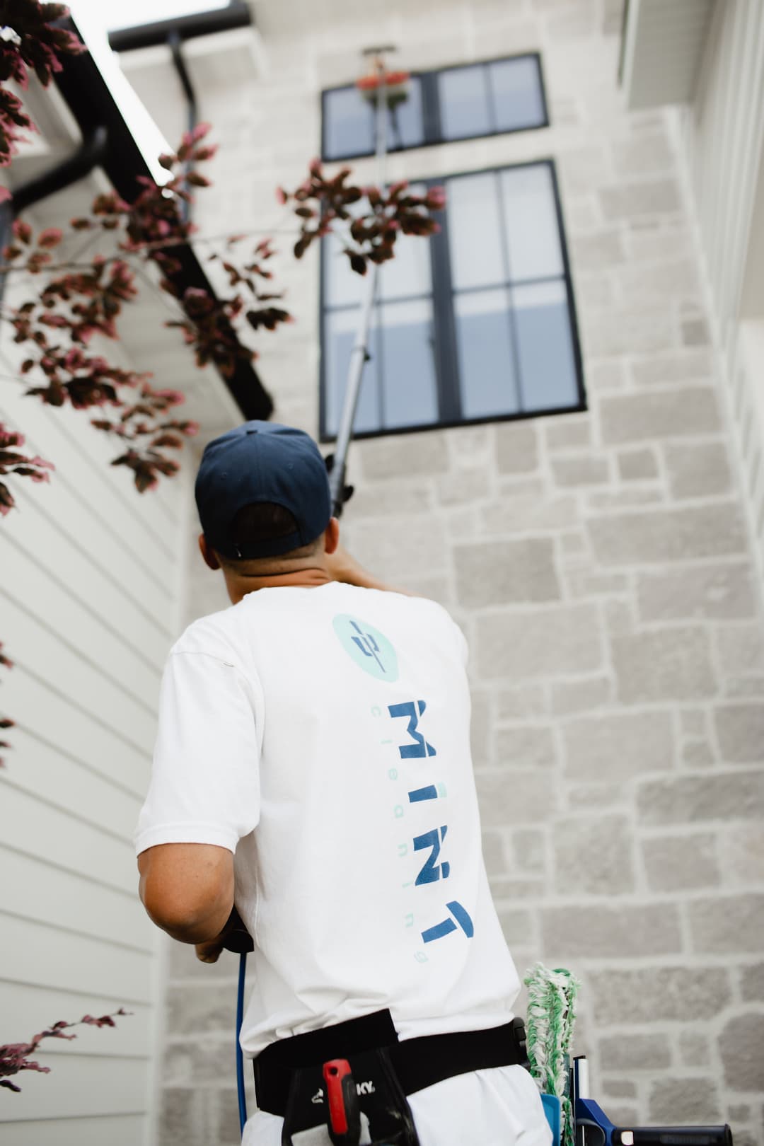 Person using a pole pruner to trim tree branches near a modern home exterior.