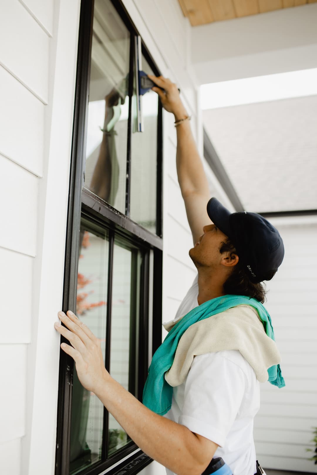 Man cleaning exterior window of a modern home, using a squeegee and wearing a blue cap.