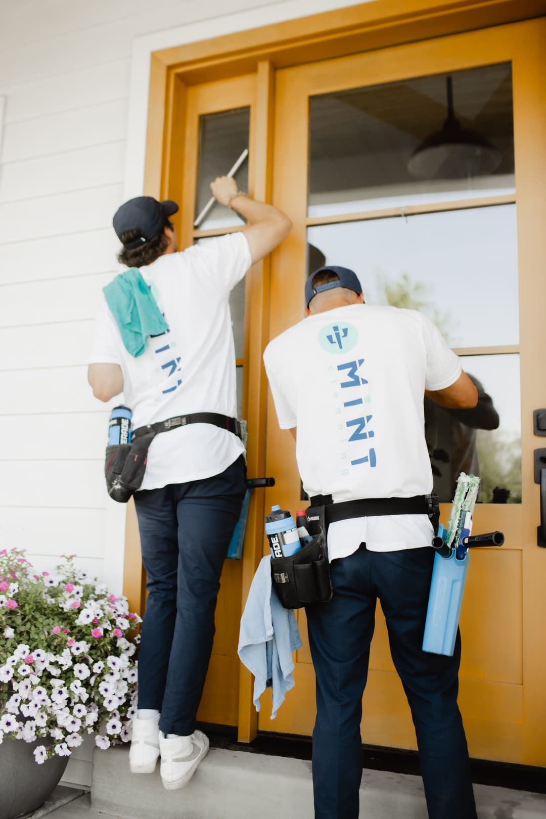 Two window cleaners in branded shirts cleaning glass doors with tools and supplies visible.