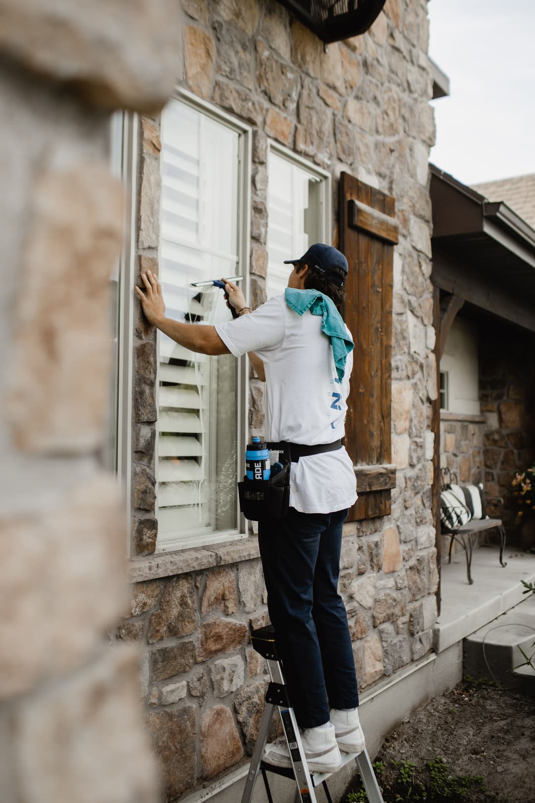 Person cleaning windows on a ladder outside a stone house, wearing a blue scarf and tool belt.