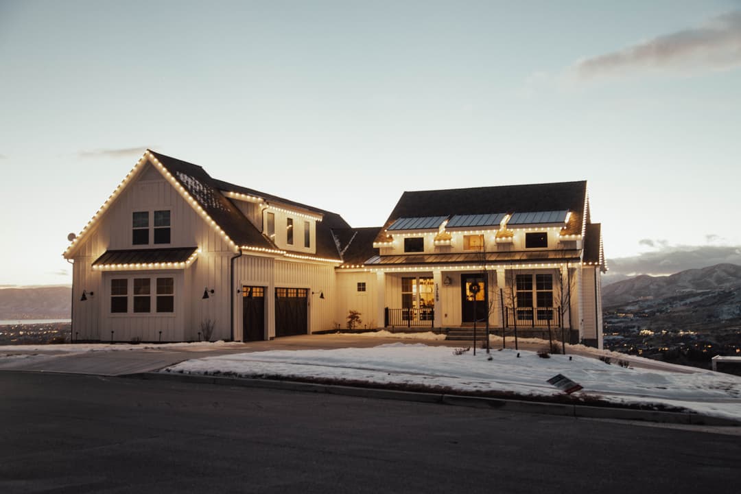 Modern white house with festive lights, snow-covered yard, and mountain backdrop at sunset.