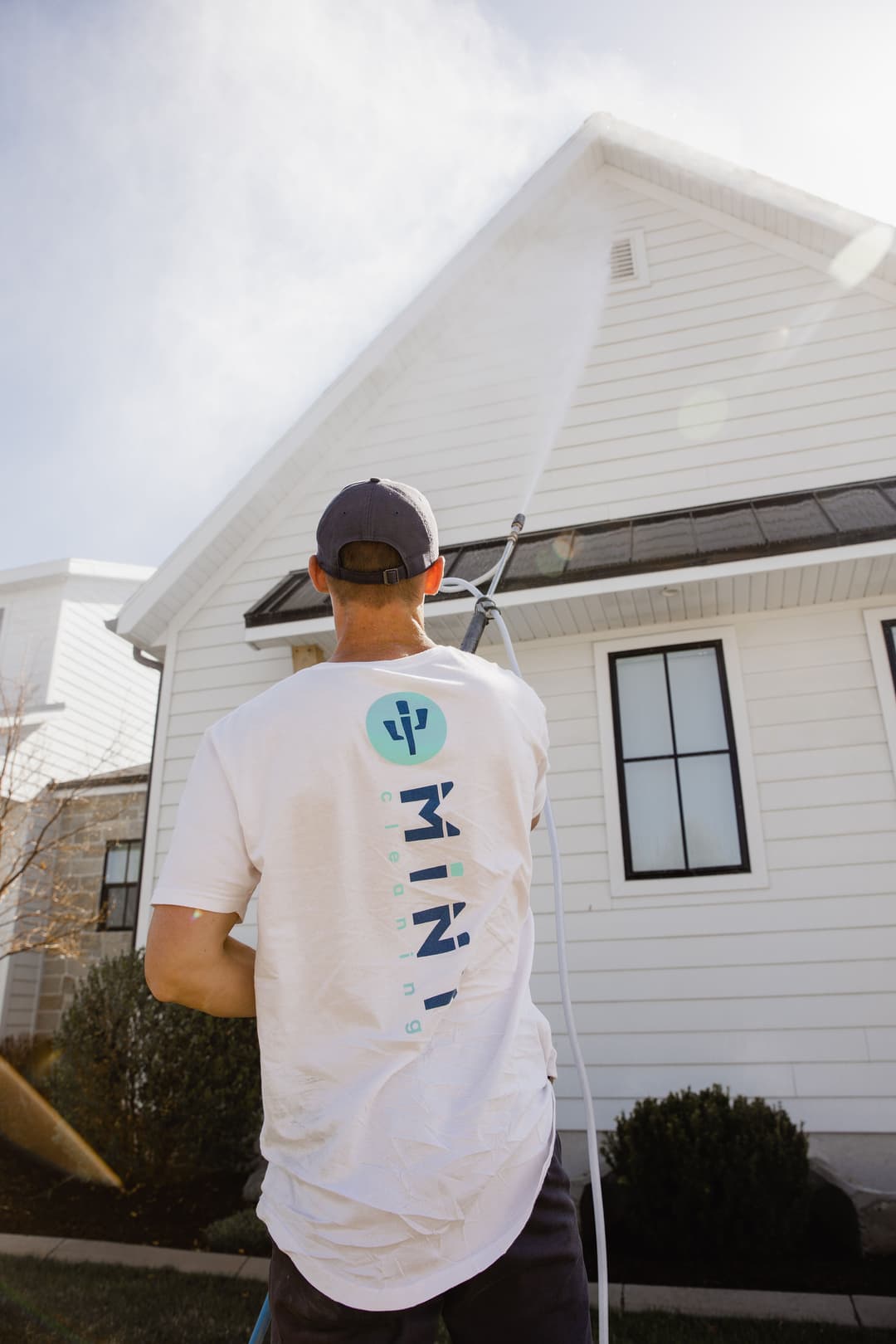 Man pressure washing a white house, wearing a branded shirt, with clear blue sky above.