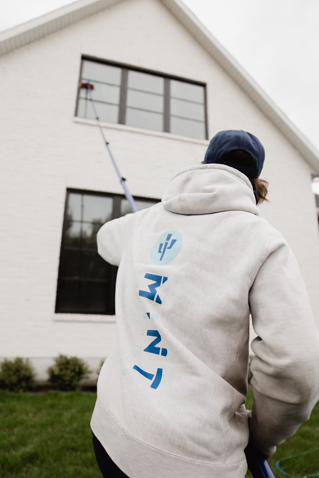 Person cleaning large windows on a house with a long reach pole, wearing a hoodie.