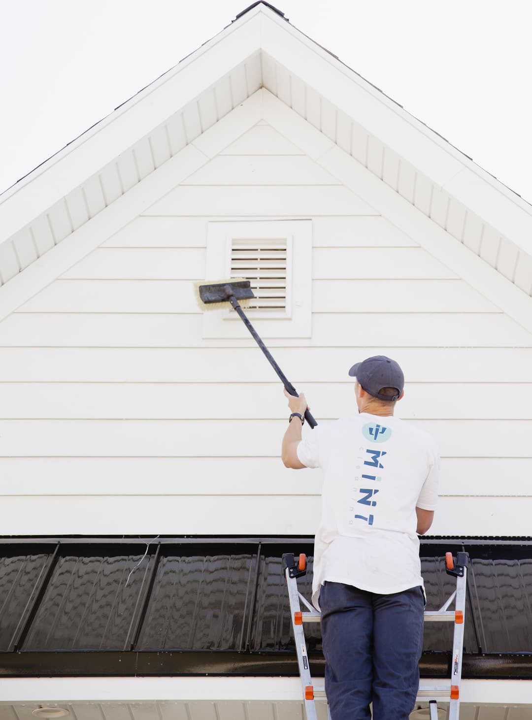 Person using a long pole to paint the upper section of a white house's exterior wall.
