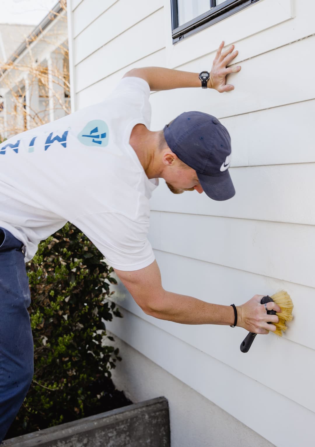 Man cleaning exterior house wall with brush, wearing a cap and casual shirt. Home maintenance.