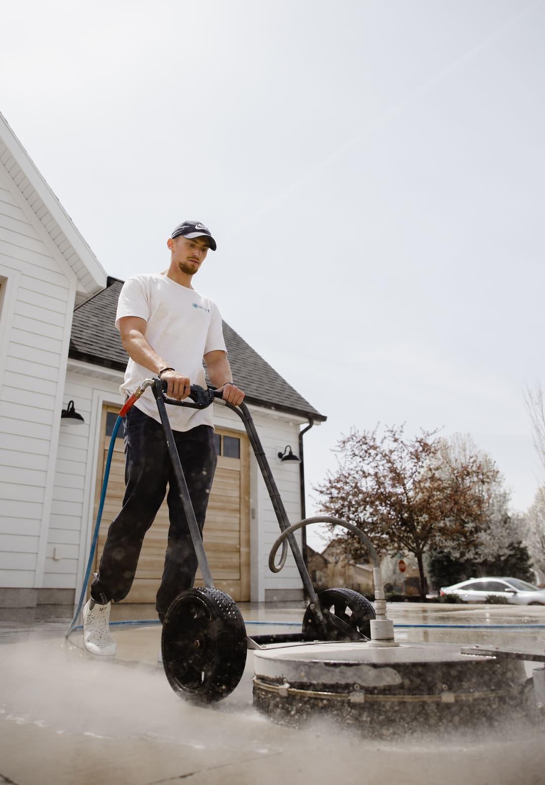 Man using a pressure washer on a driveway in front of a house on a sunny day.