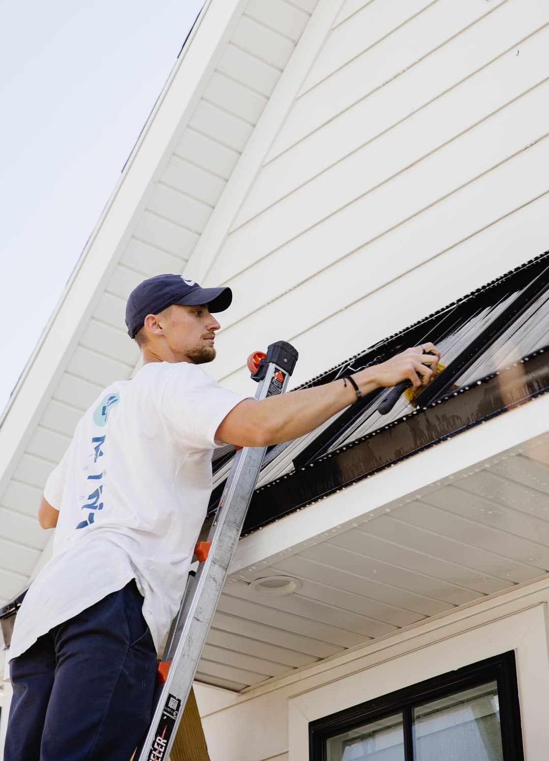 Man on ladder cleaning gutters on a white house exterior.
