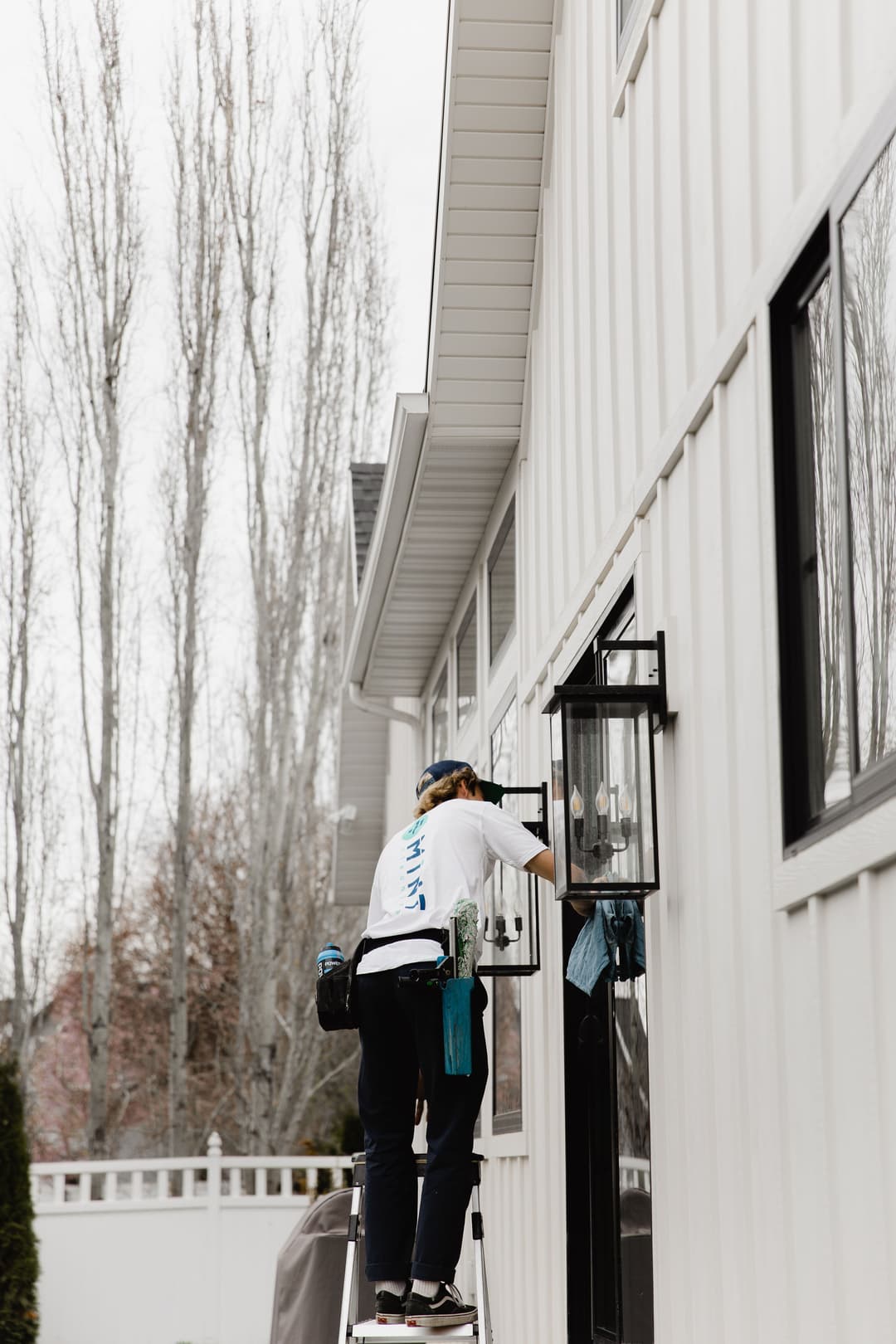 Person cleaning a window on a house using a ladder, surrounded by trees in the background.