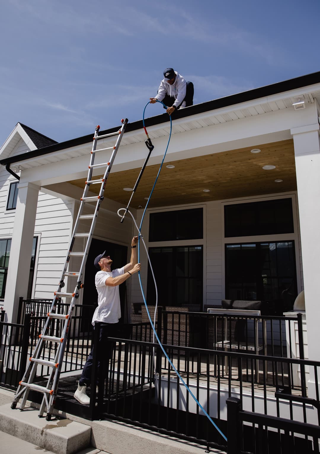 Workers using a hose to clean a roof, with a ladder and porch in the background.