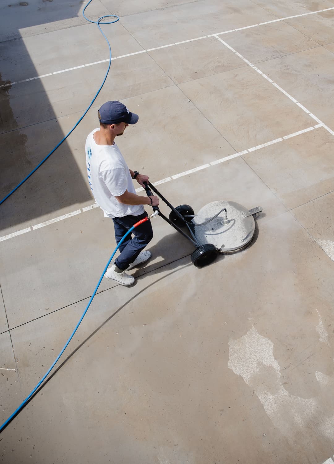 Man using a surface cleaner on a concrete floor outdoors, with a blue water hose visible.