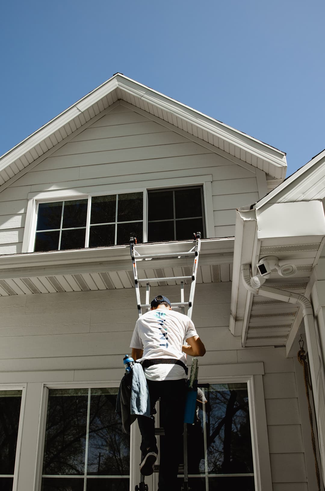 Worker cleaning upper windows of a house using a ladder on a sunny day.