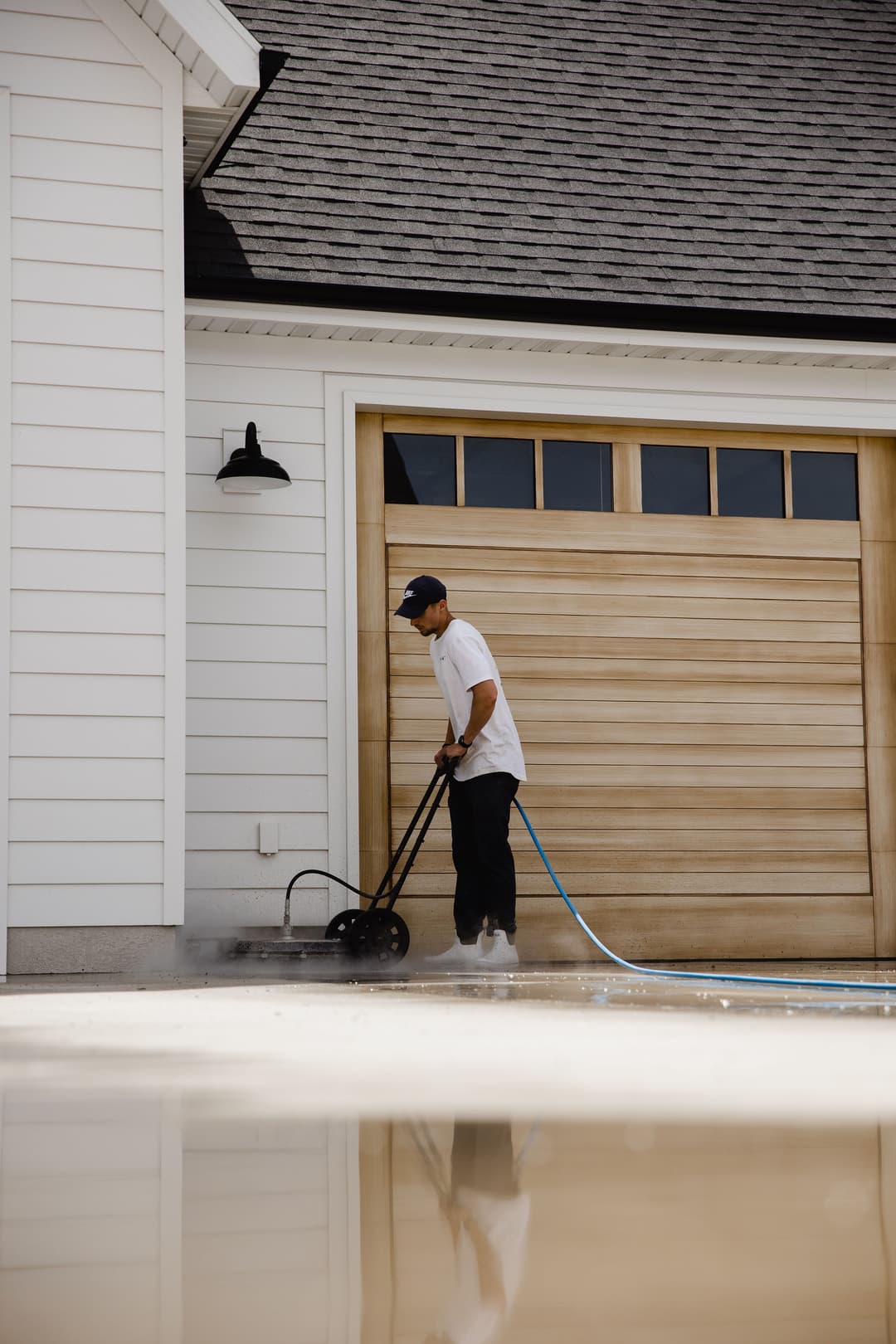 Person using a pressure washer on a driveway near a garage with a wooden door.
