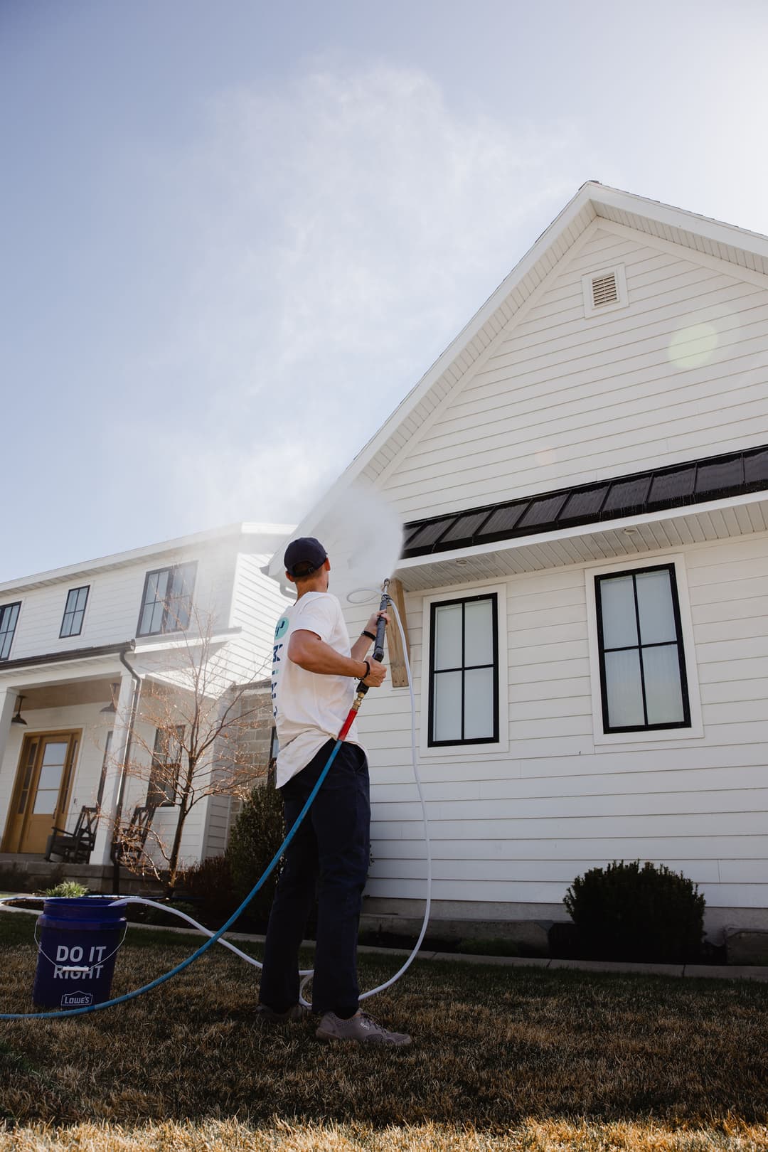 Person washing a house exterior with a spray nozzle and bucket on a sunny day.