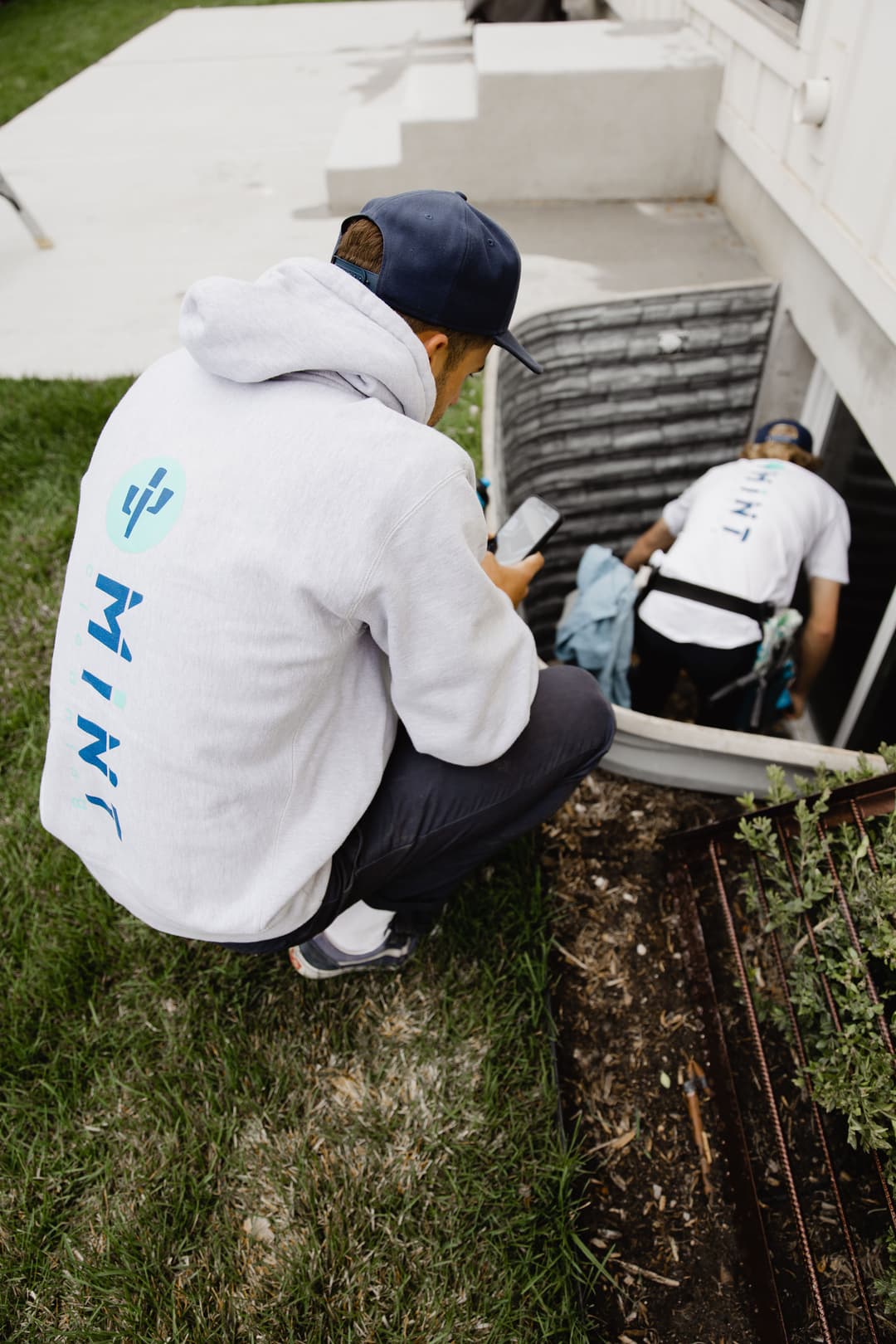 Two workers in branded hoodies performing maintenance near a basement entrance, one checking a phone.
