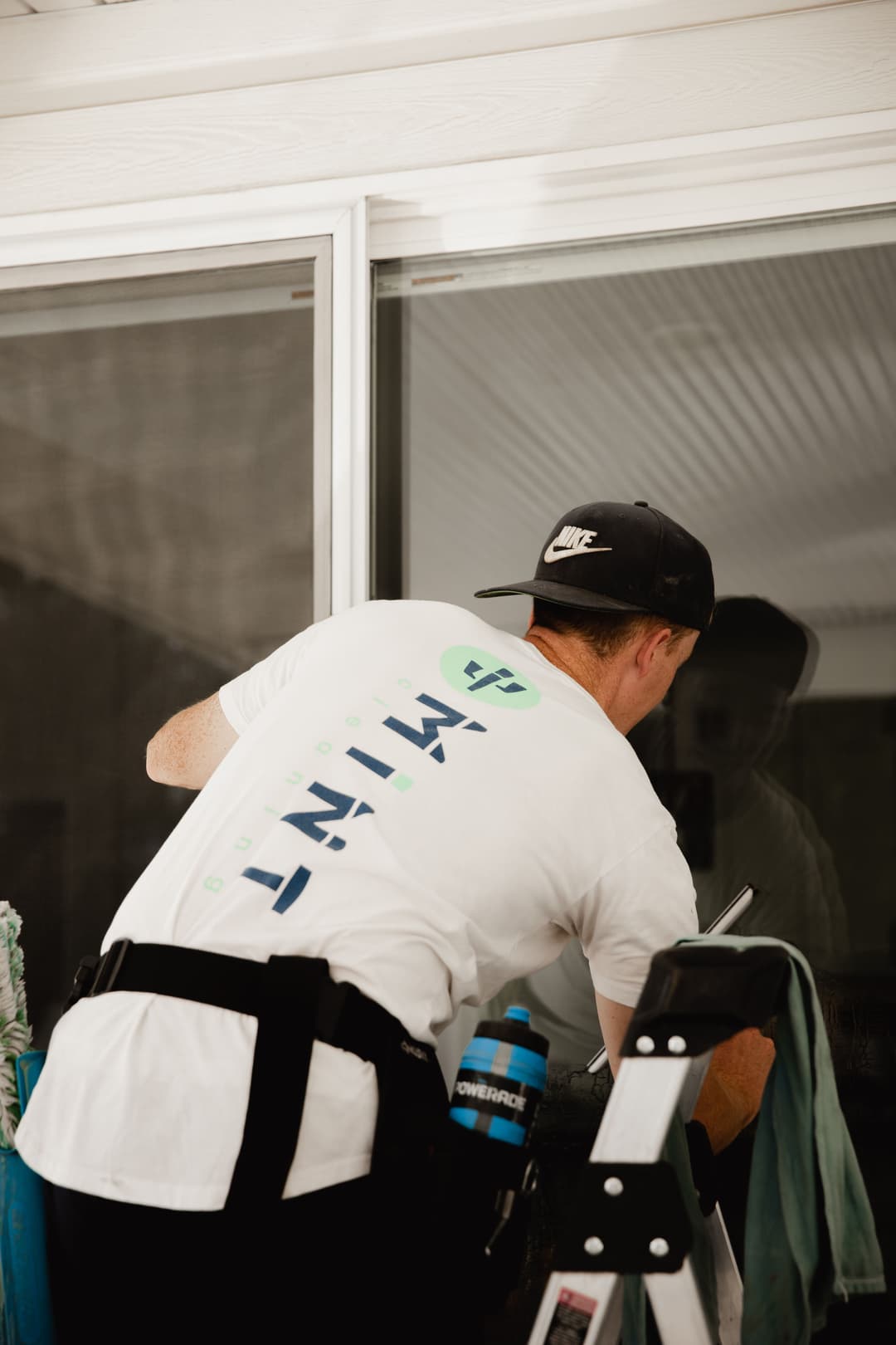 Man cleaning a glass window while using a ladder, wearing a branded T-shirt and cap.