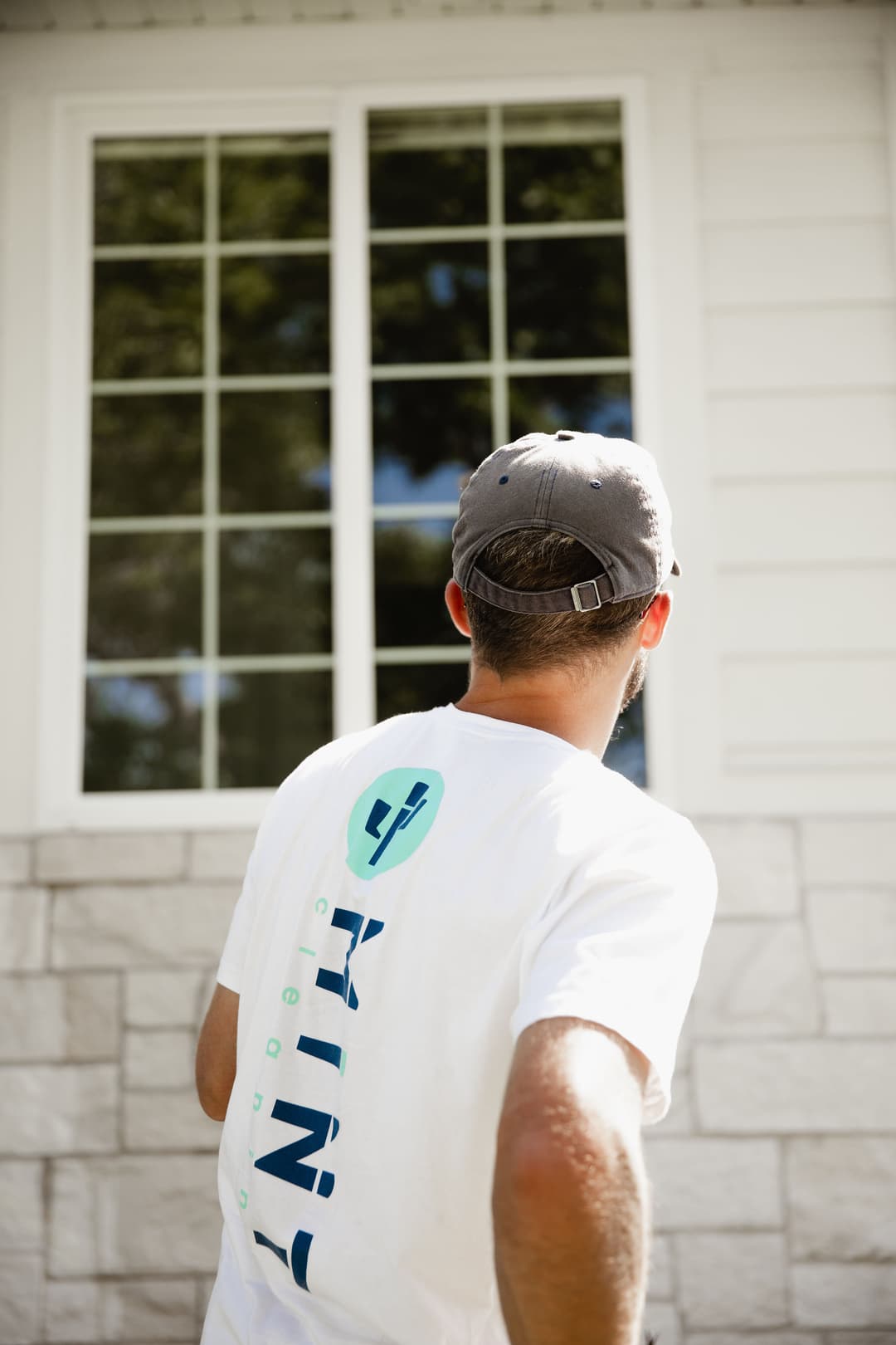 Man in a white shirt with logo, inspecting a house window outdoors.
