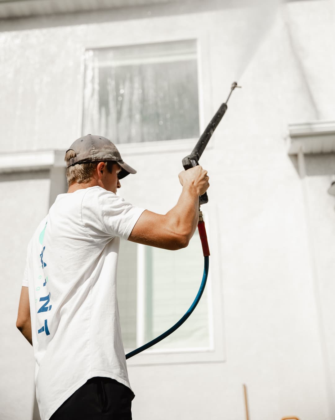 Man pressure washing a house exterior on a sunny day, wearing a cap and a white shirt.