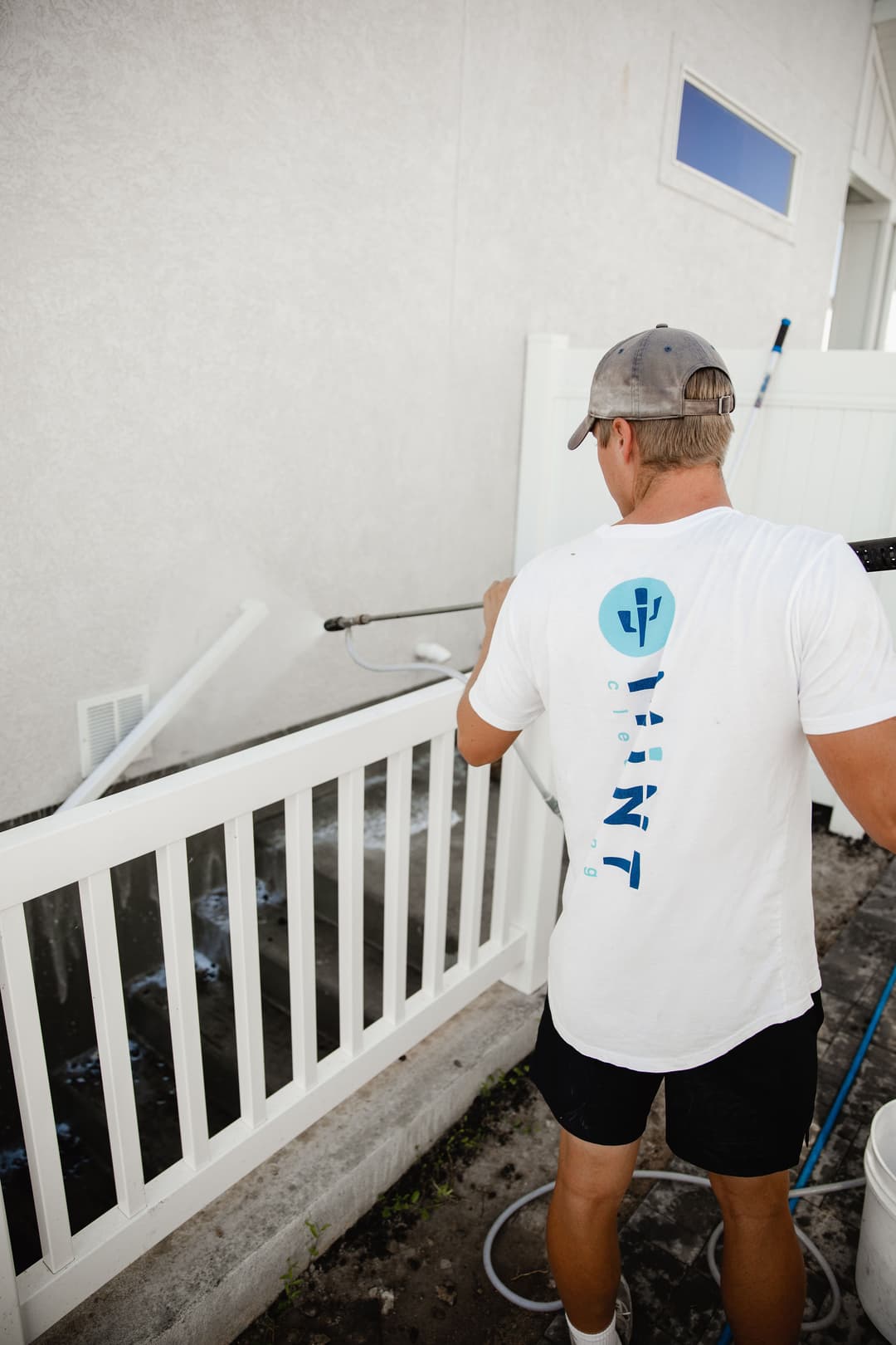 Man power washing a white fence beside a building in sunny outdoor setting.