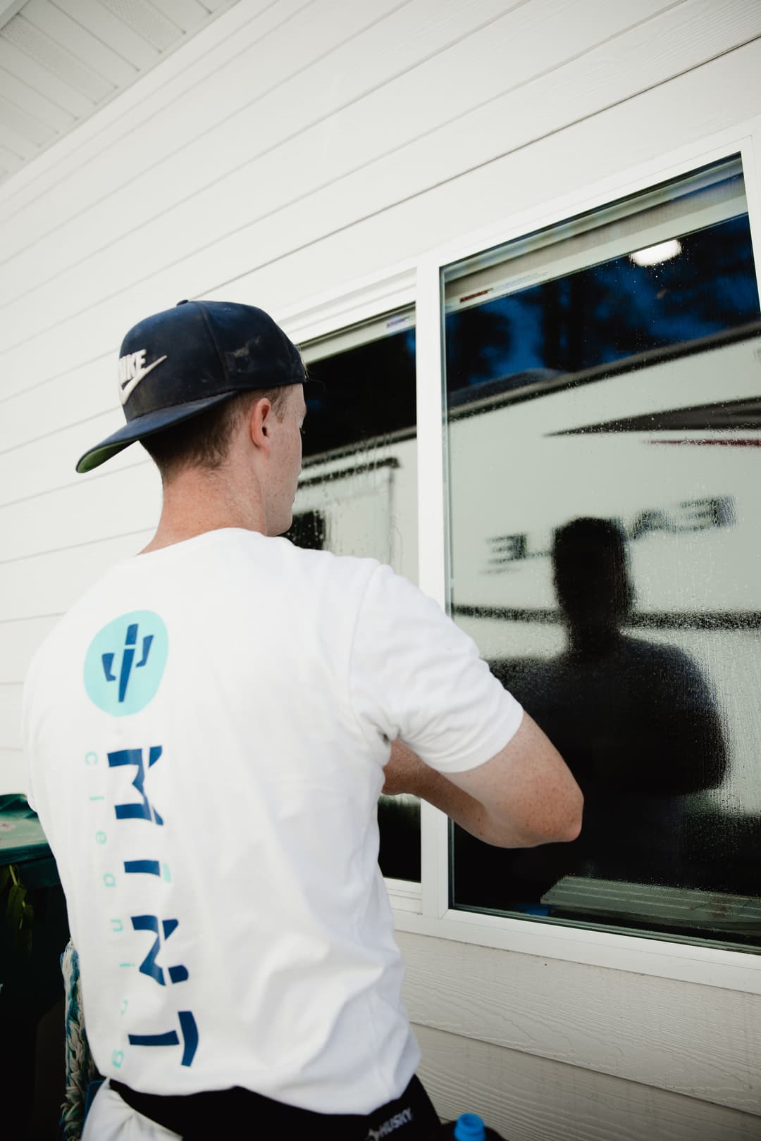 Person applying window tint to a house, wearing a branded white T-shirt and baseball cap.