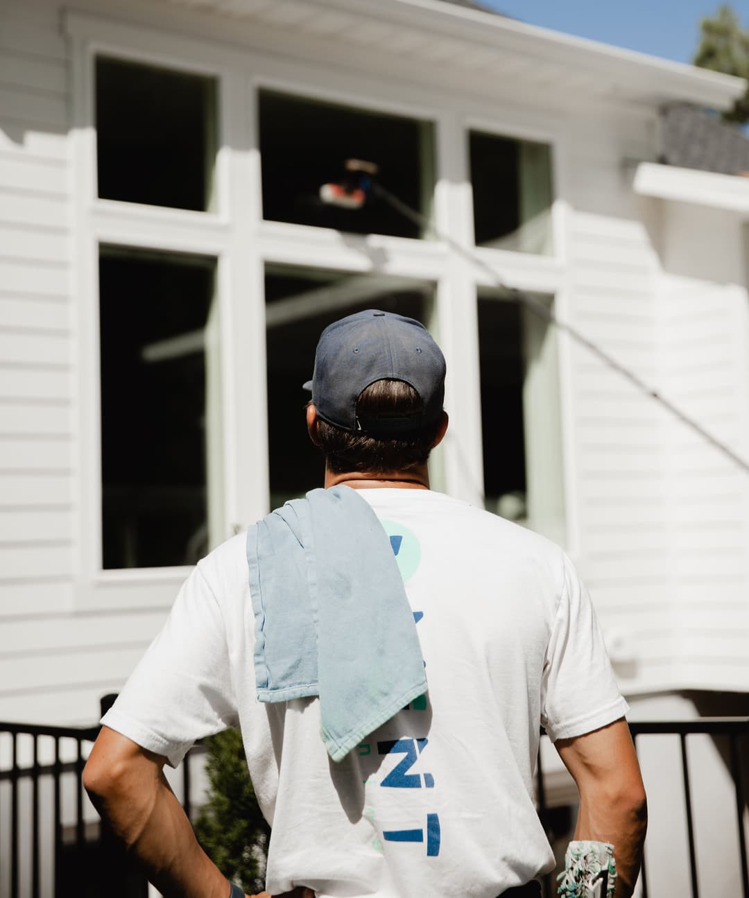 Man cleaning large windows on a house with a pole squeegee, wearing a cap and carrying a towel.