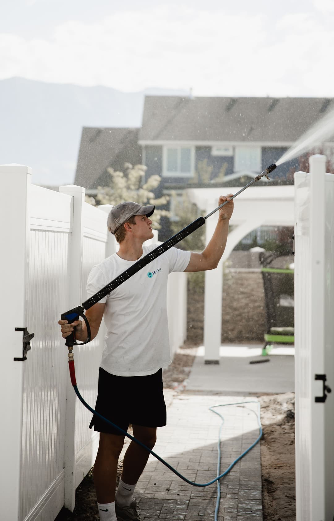 Man using a pressure washer to clean a white fence in a residential yard.