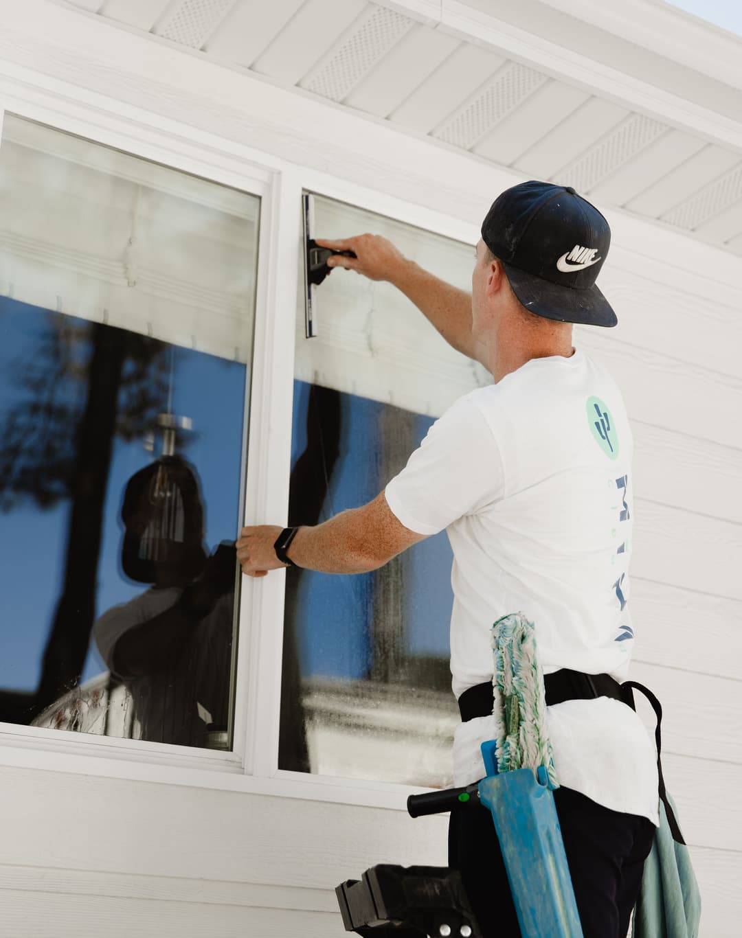 Man cleaning a window with a squeegee while using a ladder, wearing a cap and t-shirt.