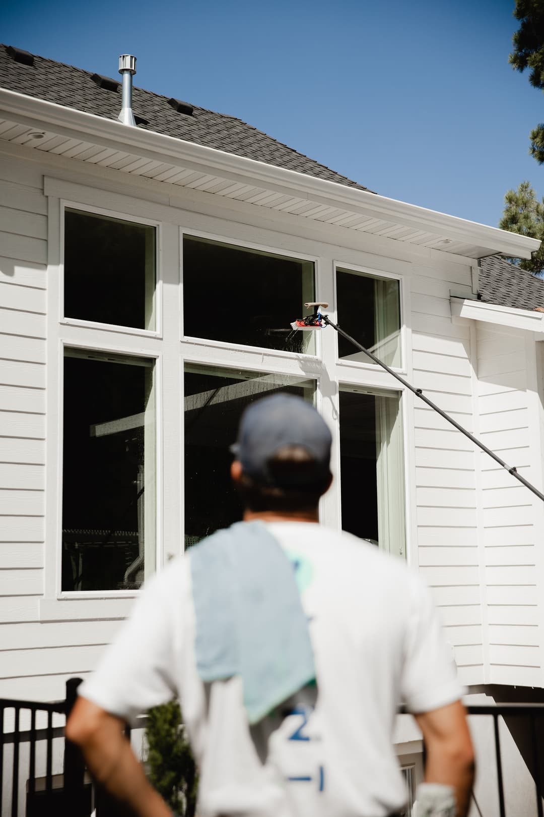 Person cleaning large windows of a modern white house with a long pole and squeegee.