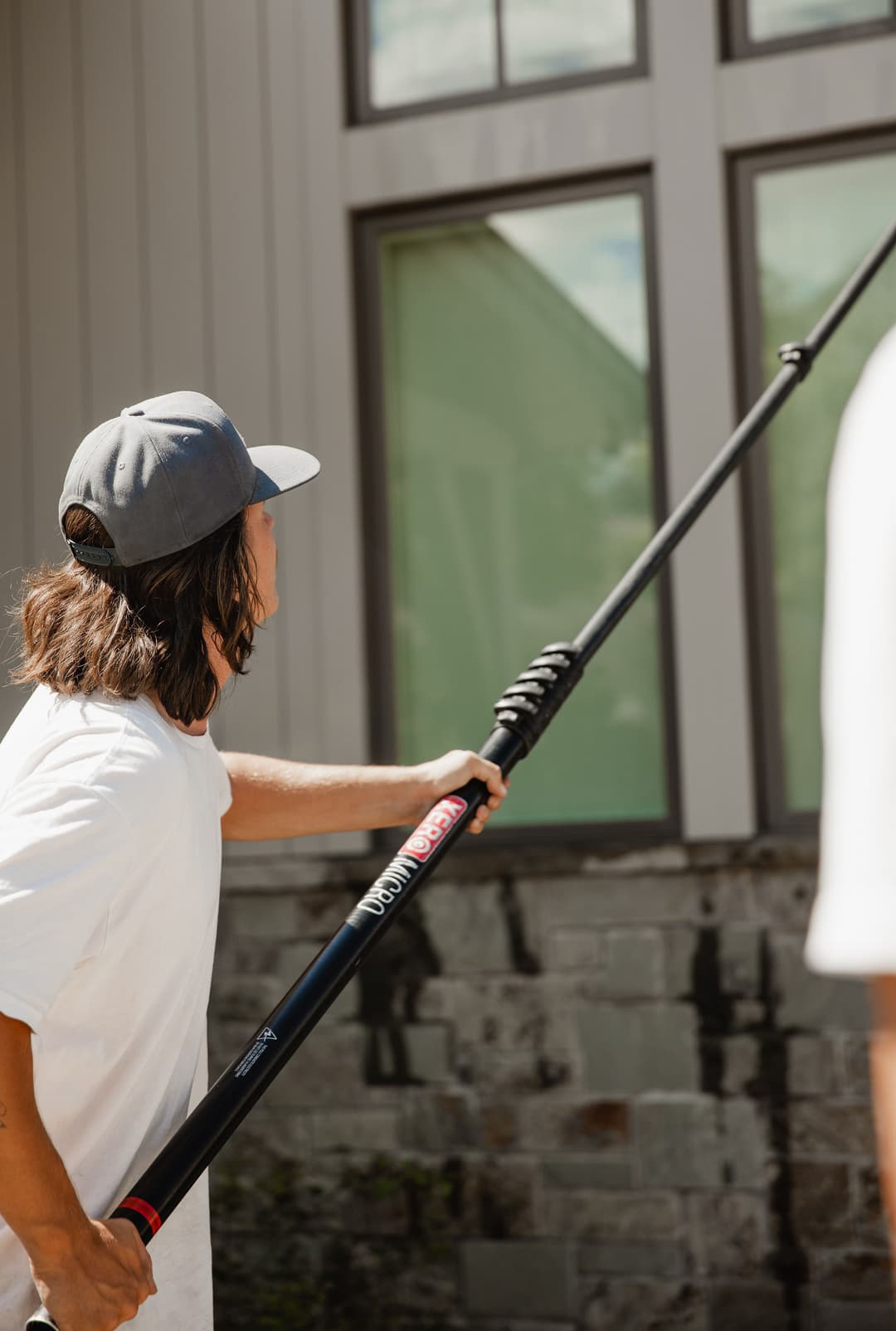 Person cleaning windows with a pole, wearing a cap and casual clothing, sunny outdoor setting.
