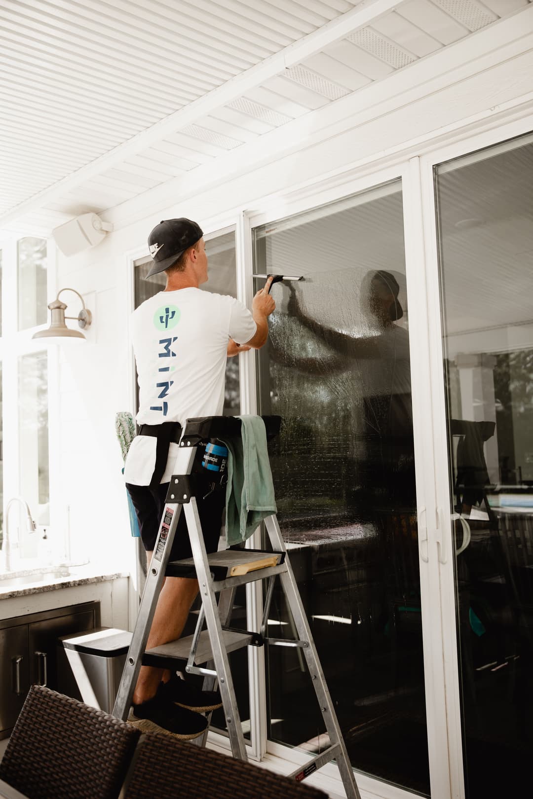 Window cleaner on ladder using squeegee on large glass window in bright, modern home.