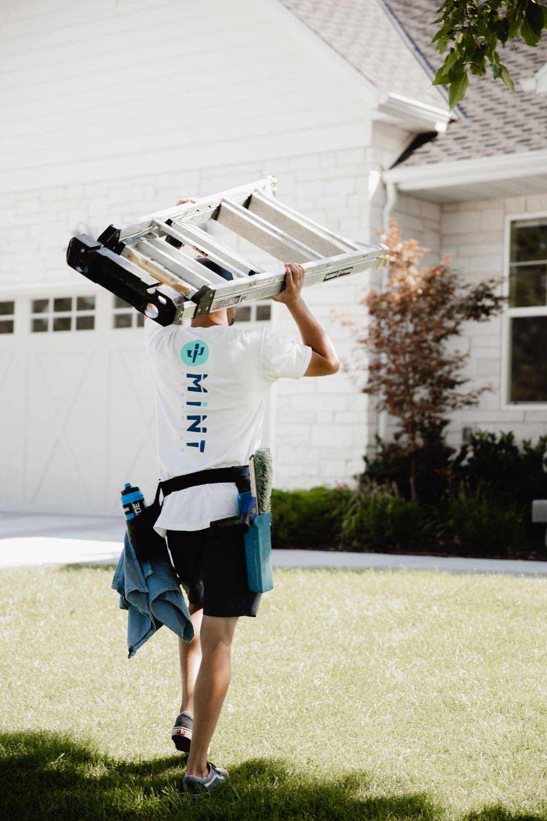 Person in a white shirt carries a ladder while wearing a tool belt in a residential yard.