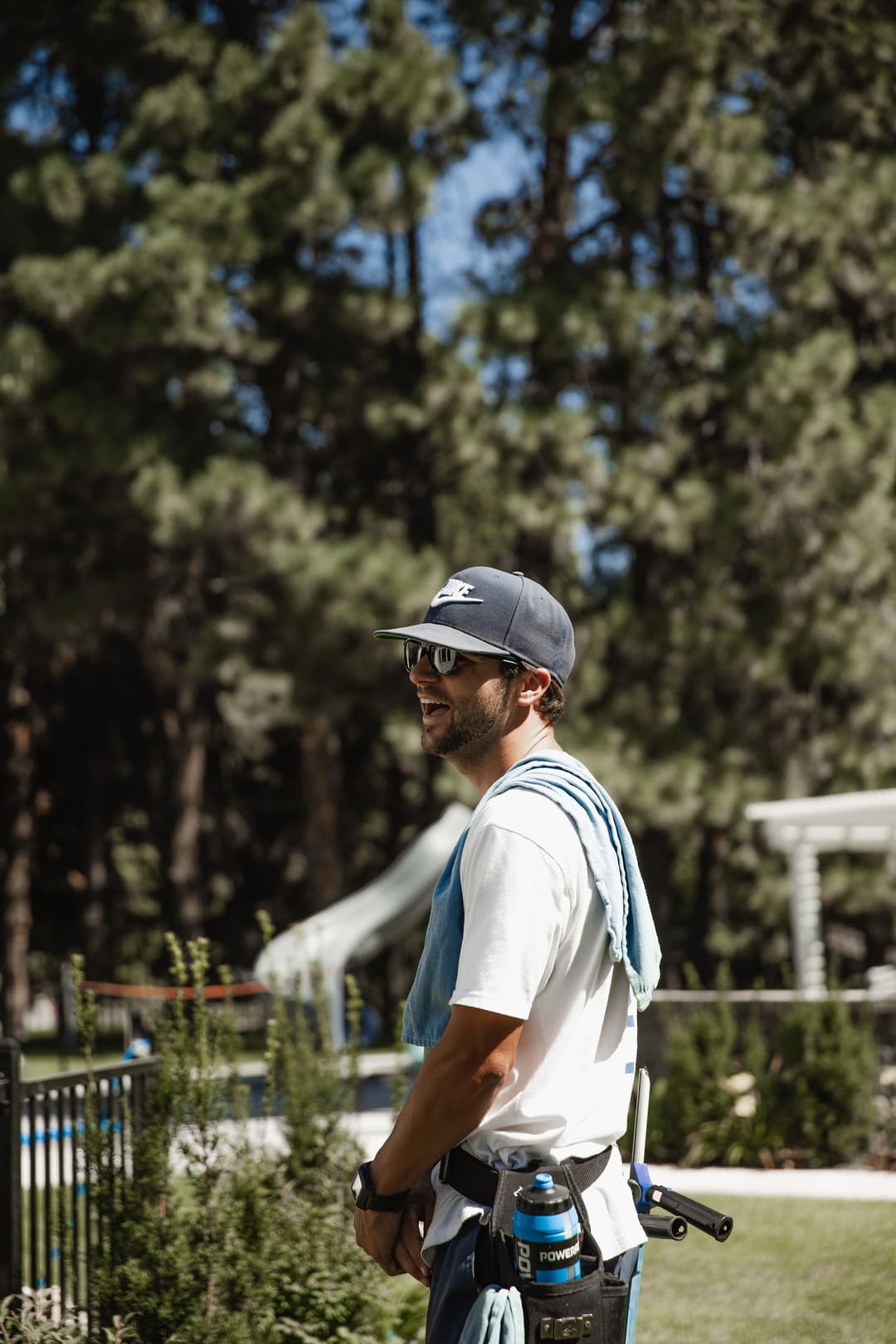 Man in baseball cap and sunglasses, smiling, with a towel and tools in a landscaped garden.