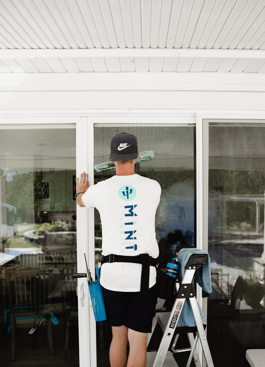 Person cleaning a large glass door with a squeegee, wearing a branded T-shirt and cap.