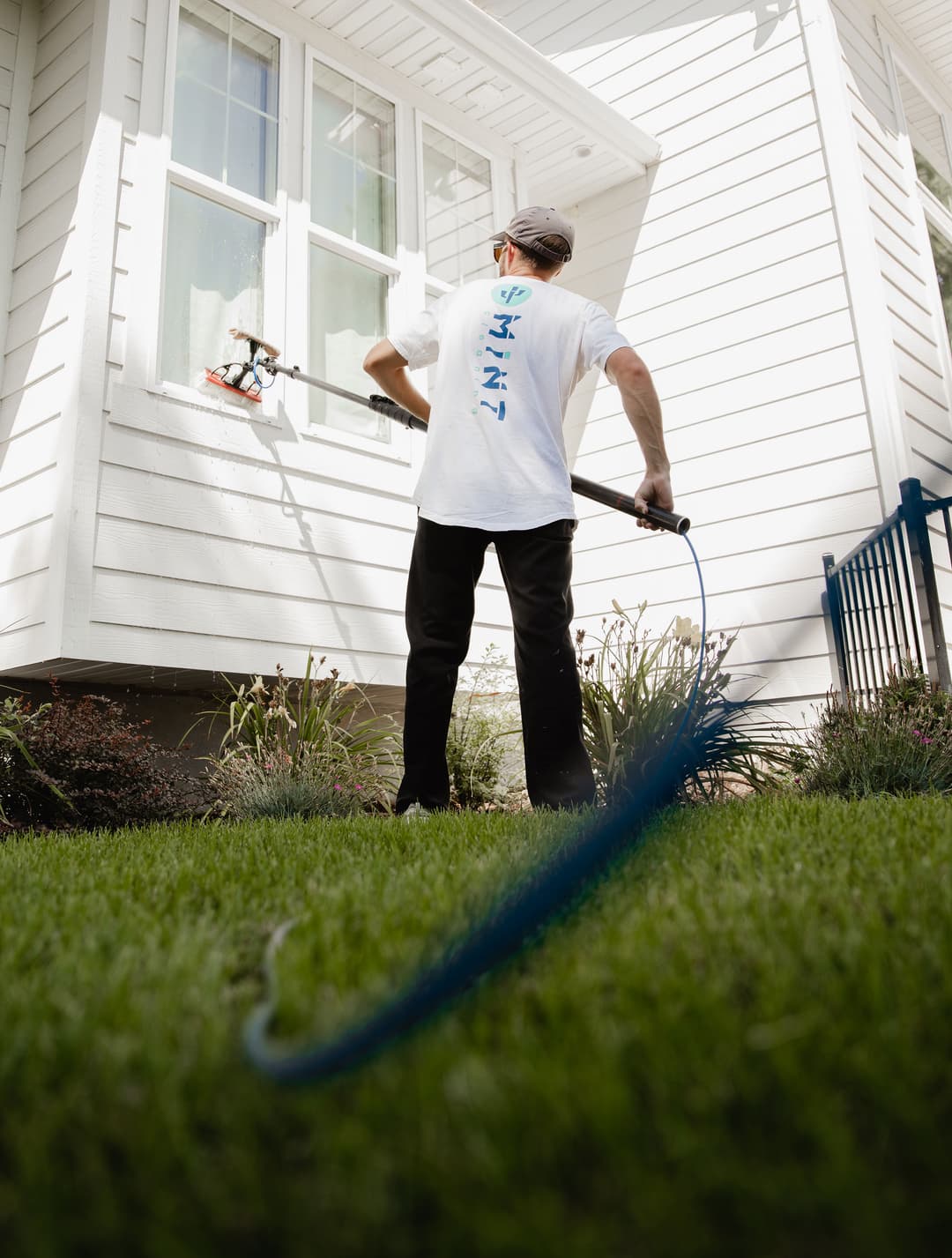 Person pressure washing a house exterior with a blue hose on a sunny day. Green lawn visible.