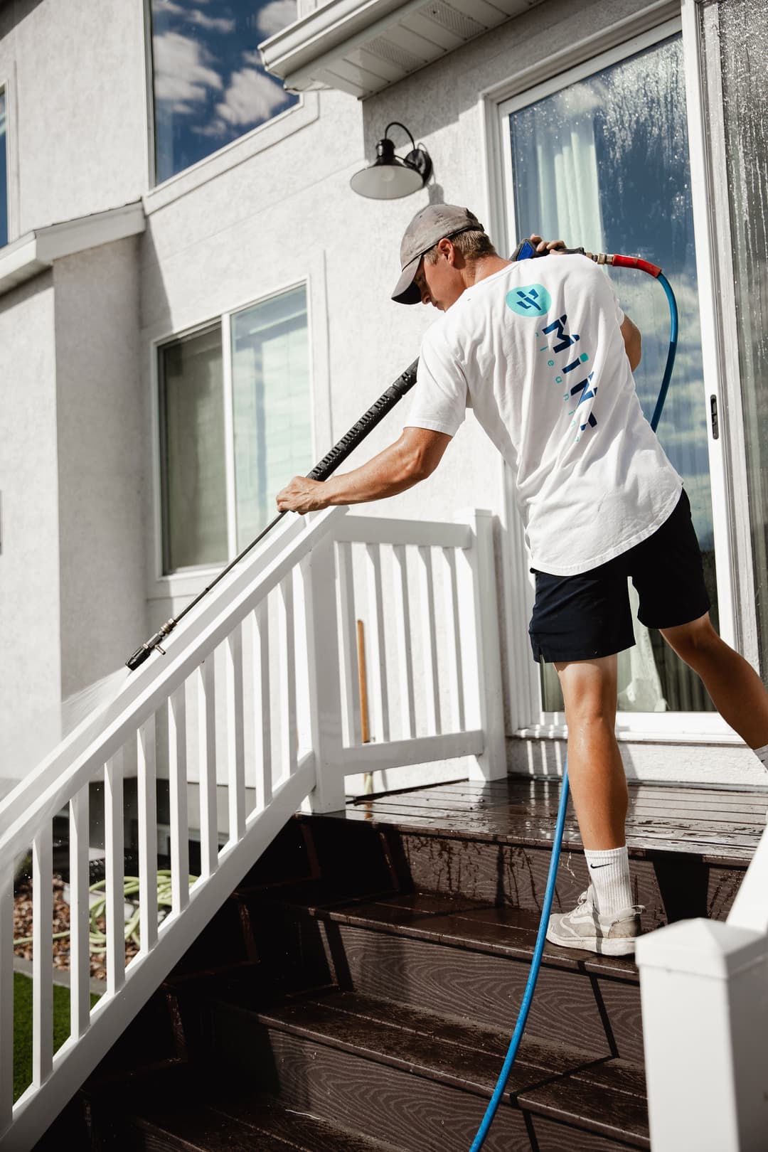 Person power washing wooden stairs of a home, wearing a white shirt and cap, with a hose.