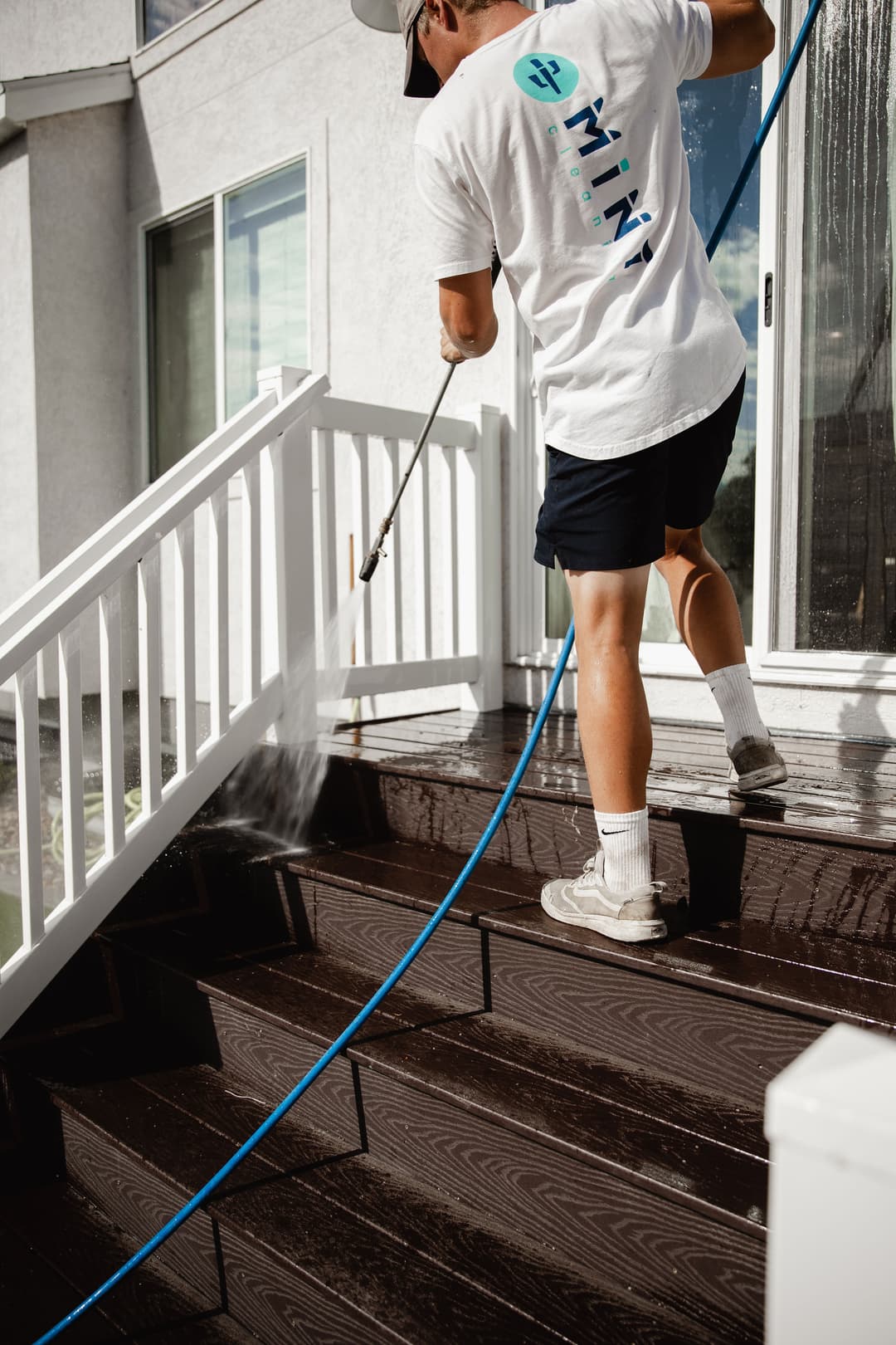 Person pressure washing a wooden deck with a hose, wearing a white shirt and shorts.