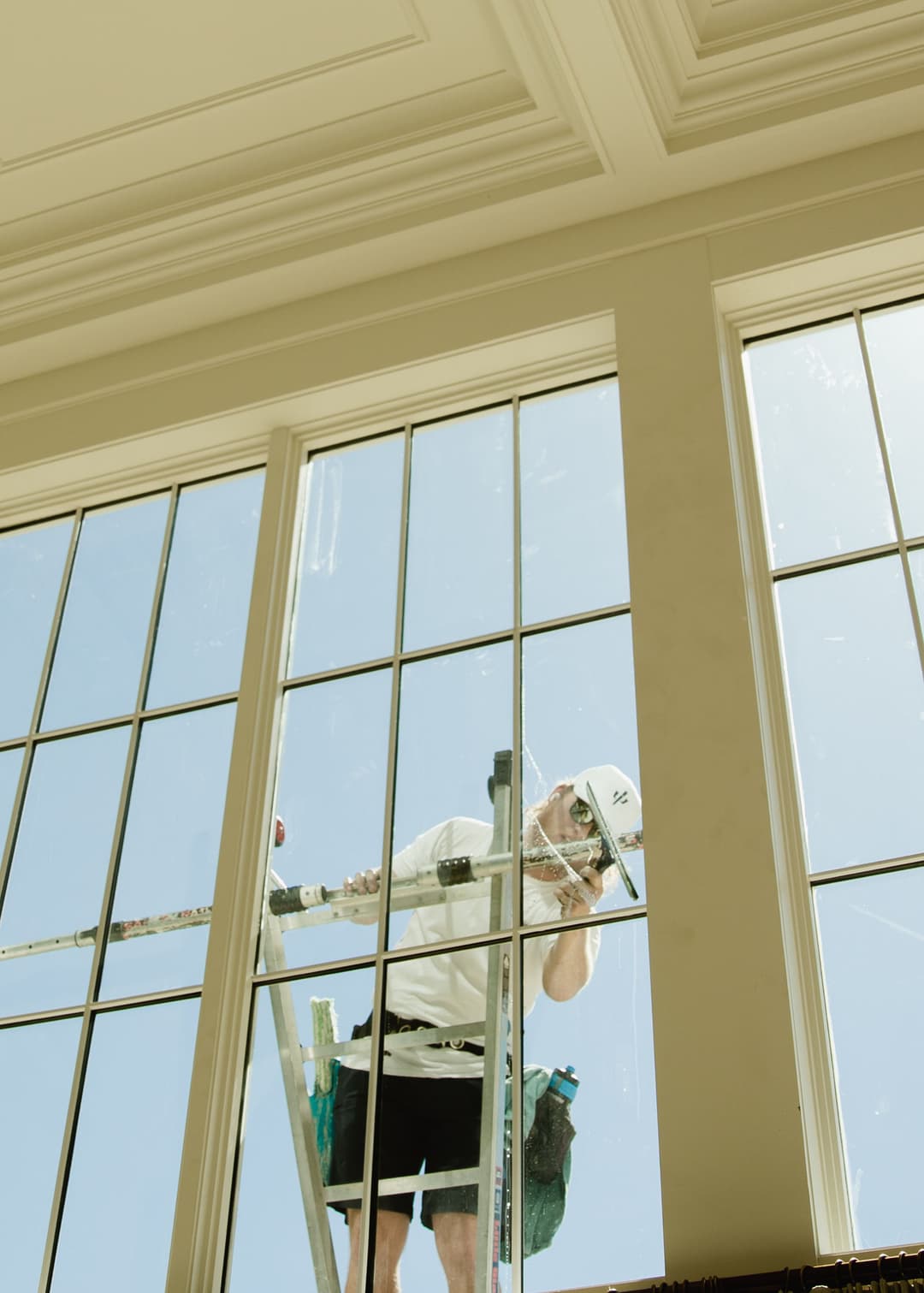 Window cleaner on a ladder, washing large pane glass windows in a sunny interior setting.