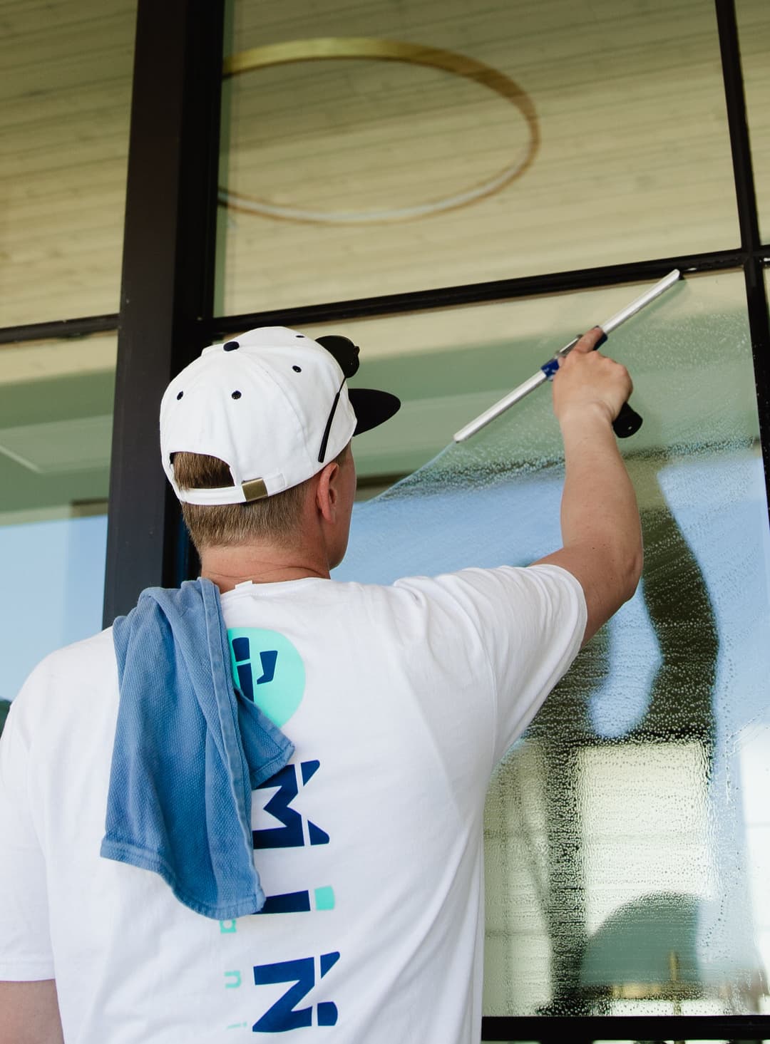 Person cleaning a large window with a squeegee, wearing a white cap and t-shirt.