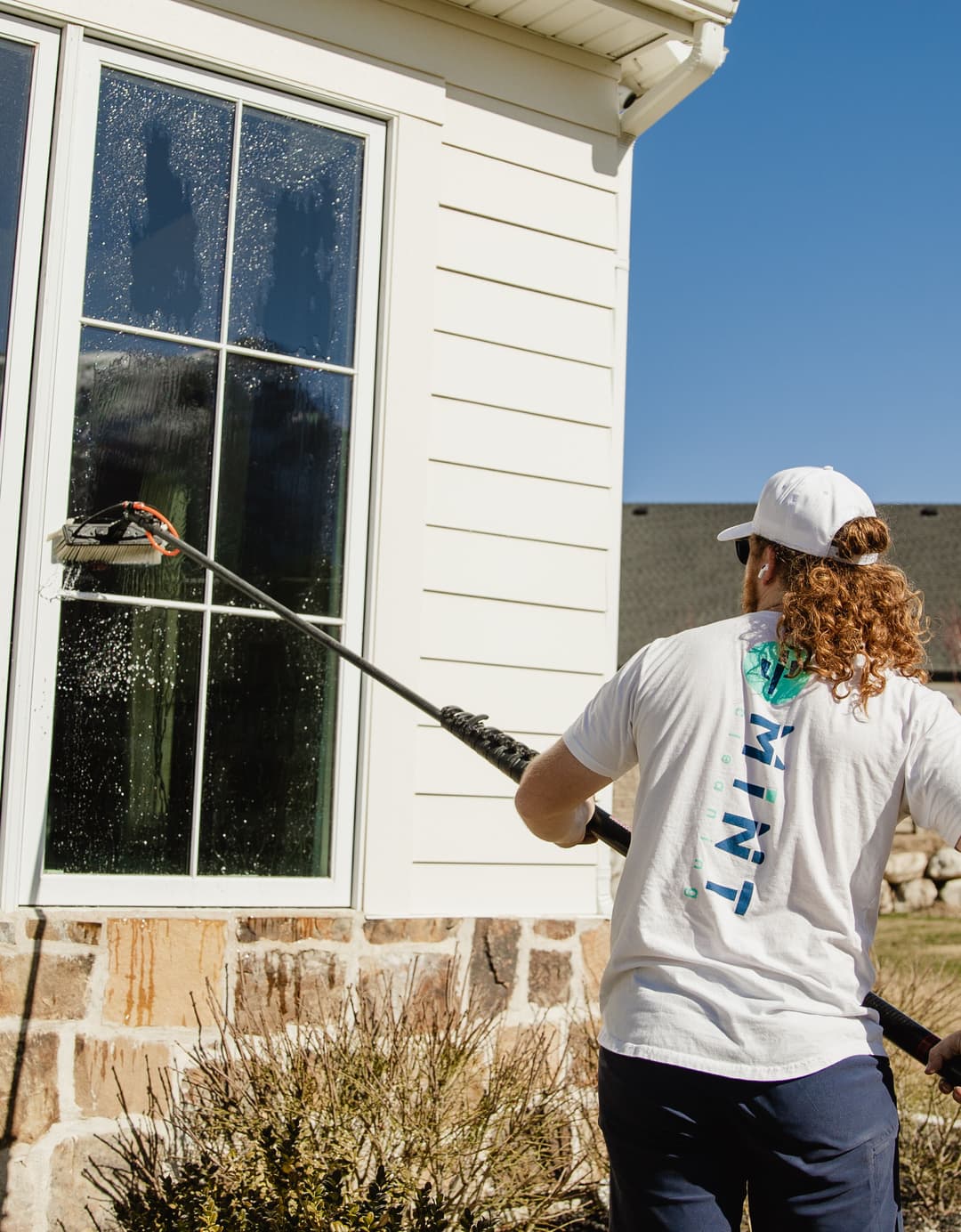 Person cleaning window with long pole, residential home exterior, blue sky background.