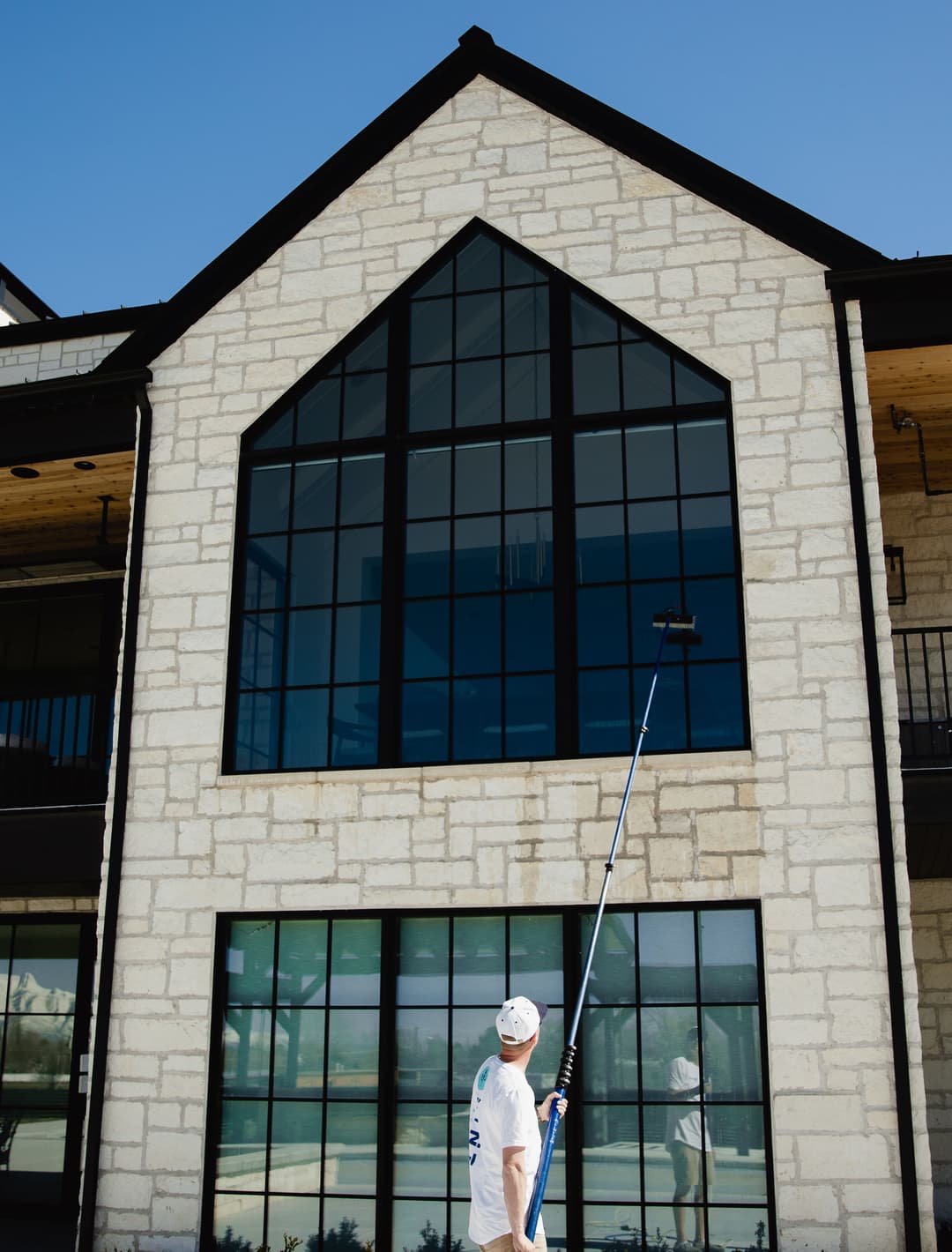 Man using a roller to clean large windows of a modern stone building under a clear blue sky.