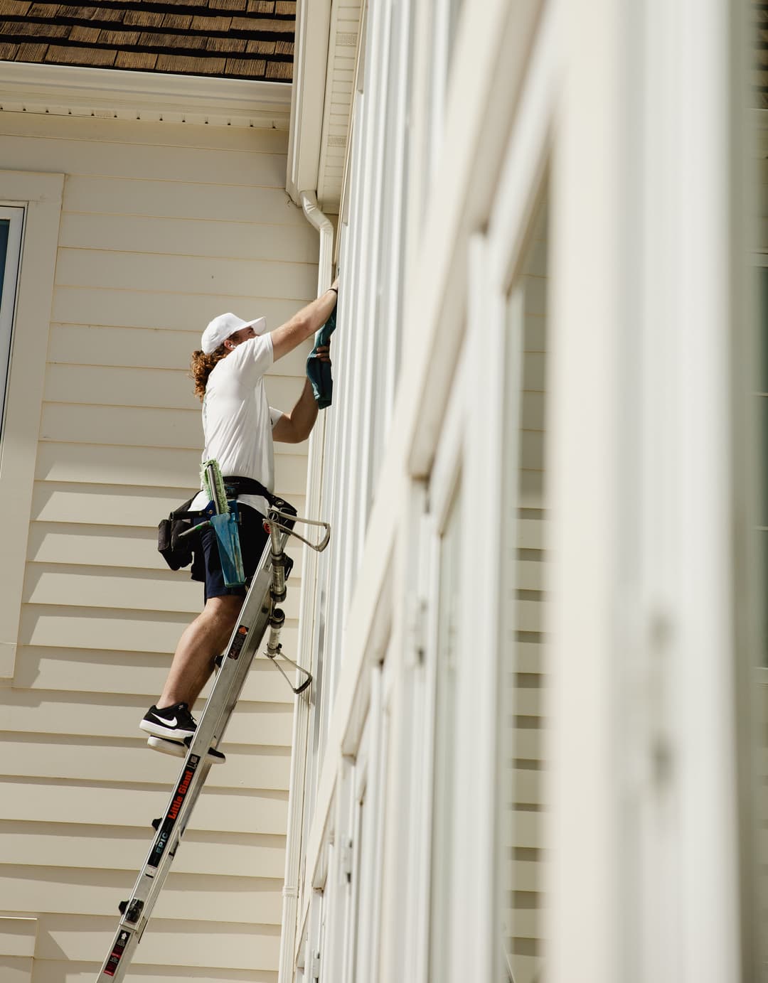 Worker on ladder performing exterior home maintenance with power tool against a white house.