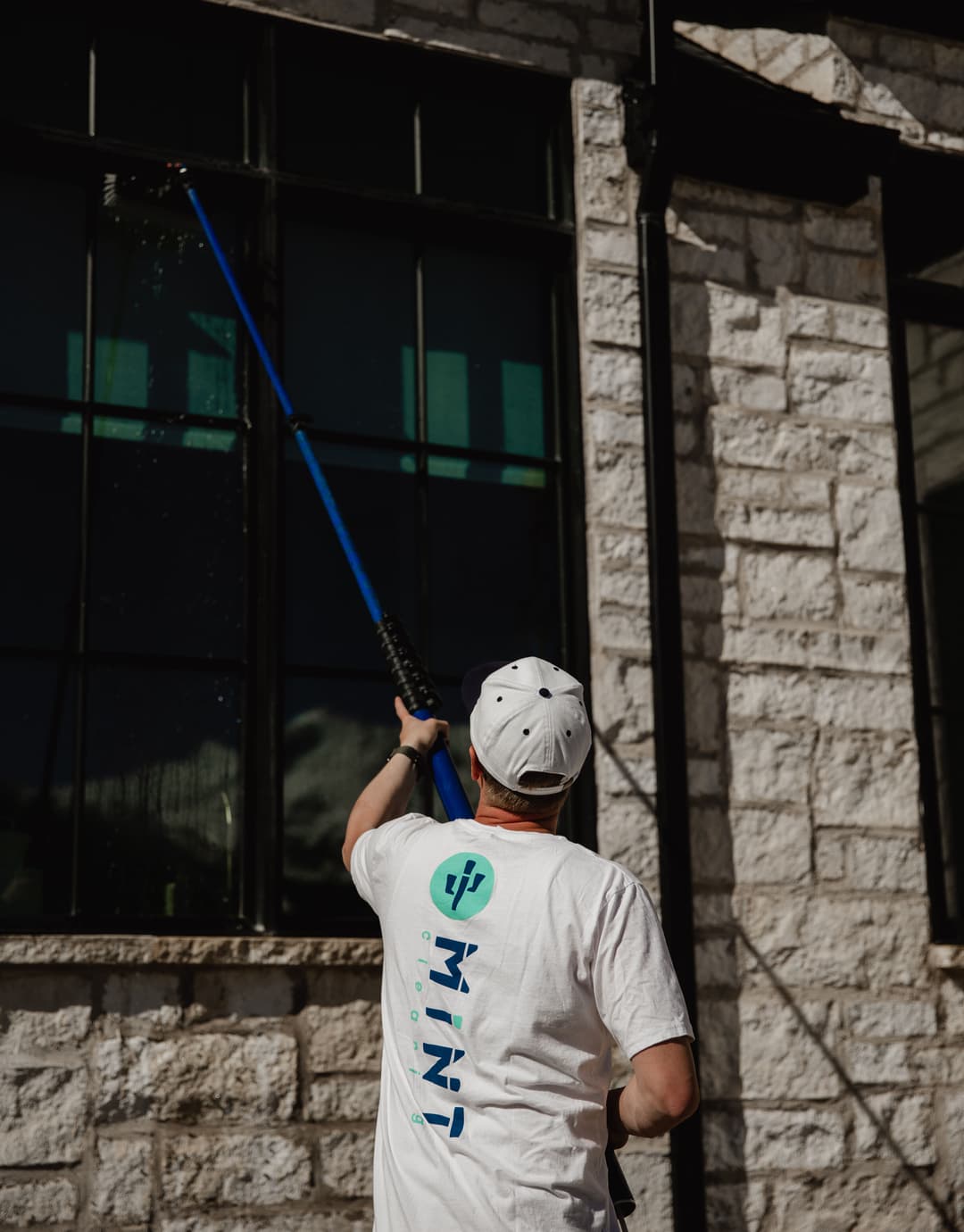 Man cleaning windows with a long pole on a stone building, wearing a white t-shirt and cap.